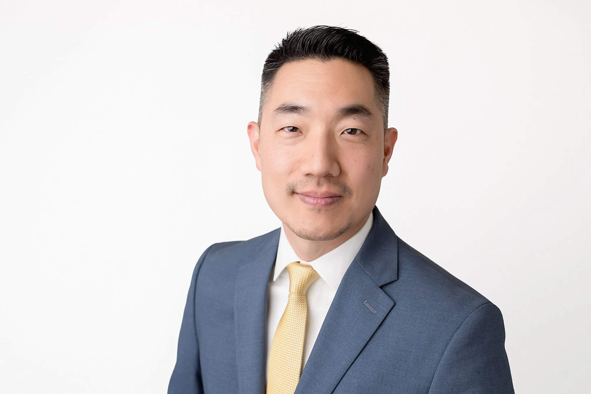 Portrait of a man in a gray suit with a yellow tie, smiling, against a plain white background during a professional headshot session with Ally and B Photography in Naperville, IL.