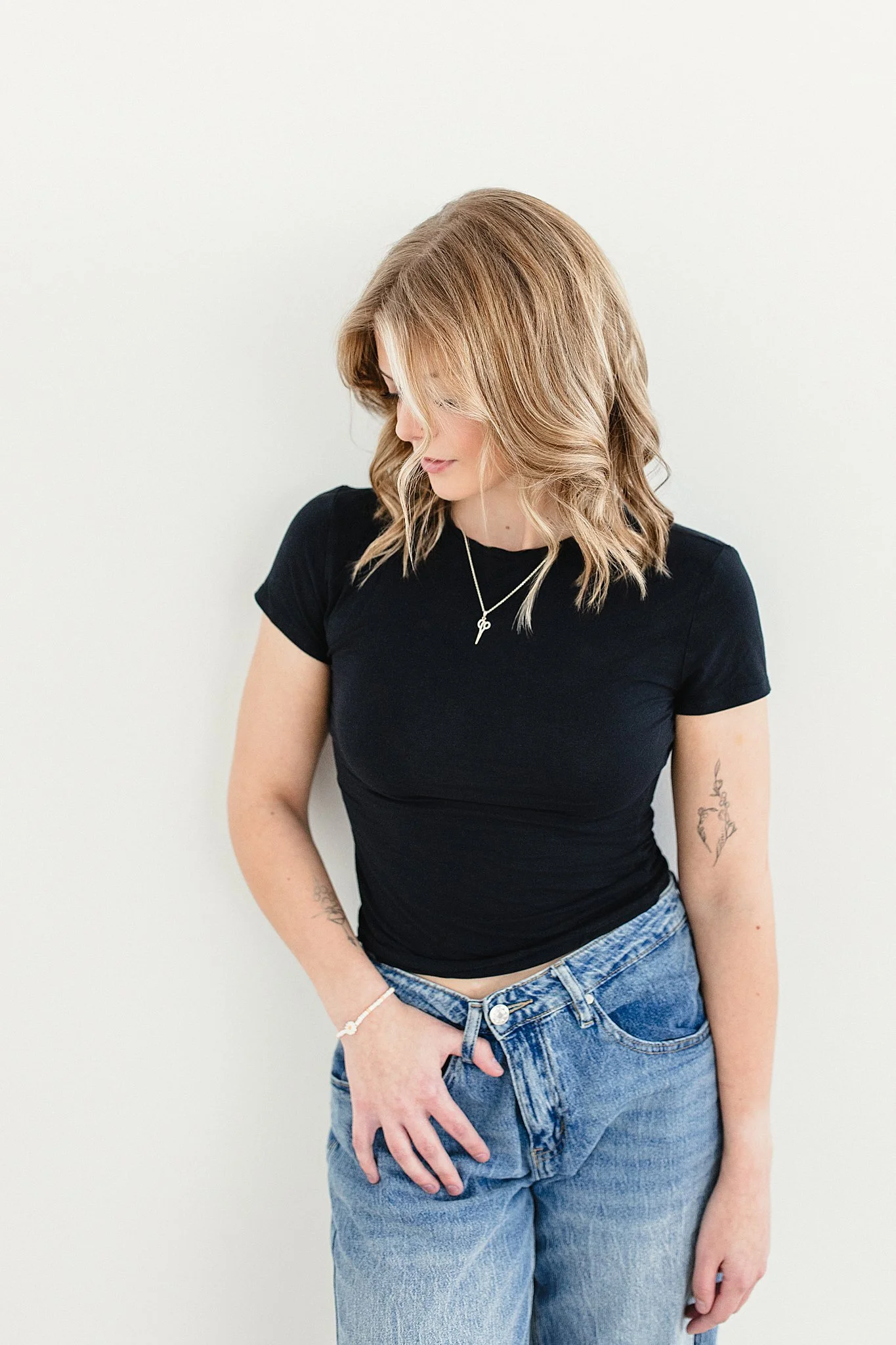 A woman with shoulder-length wavy blonde hair, wearing a black t-shirt, light blue jeans, and a silver necklace, stands against a plain white wall, looking down and to her left for a hairstylist branding session in Naperville, IL.