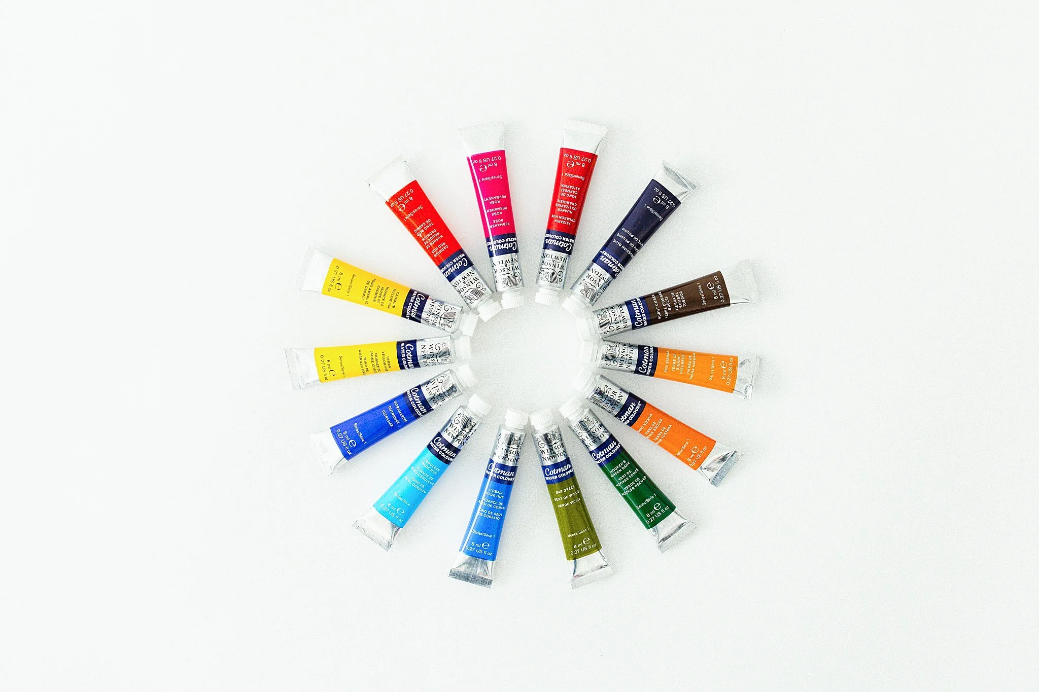 A circular arrangement of 16 watercolor tubes in various colors on a white background for product photography near me.