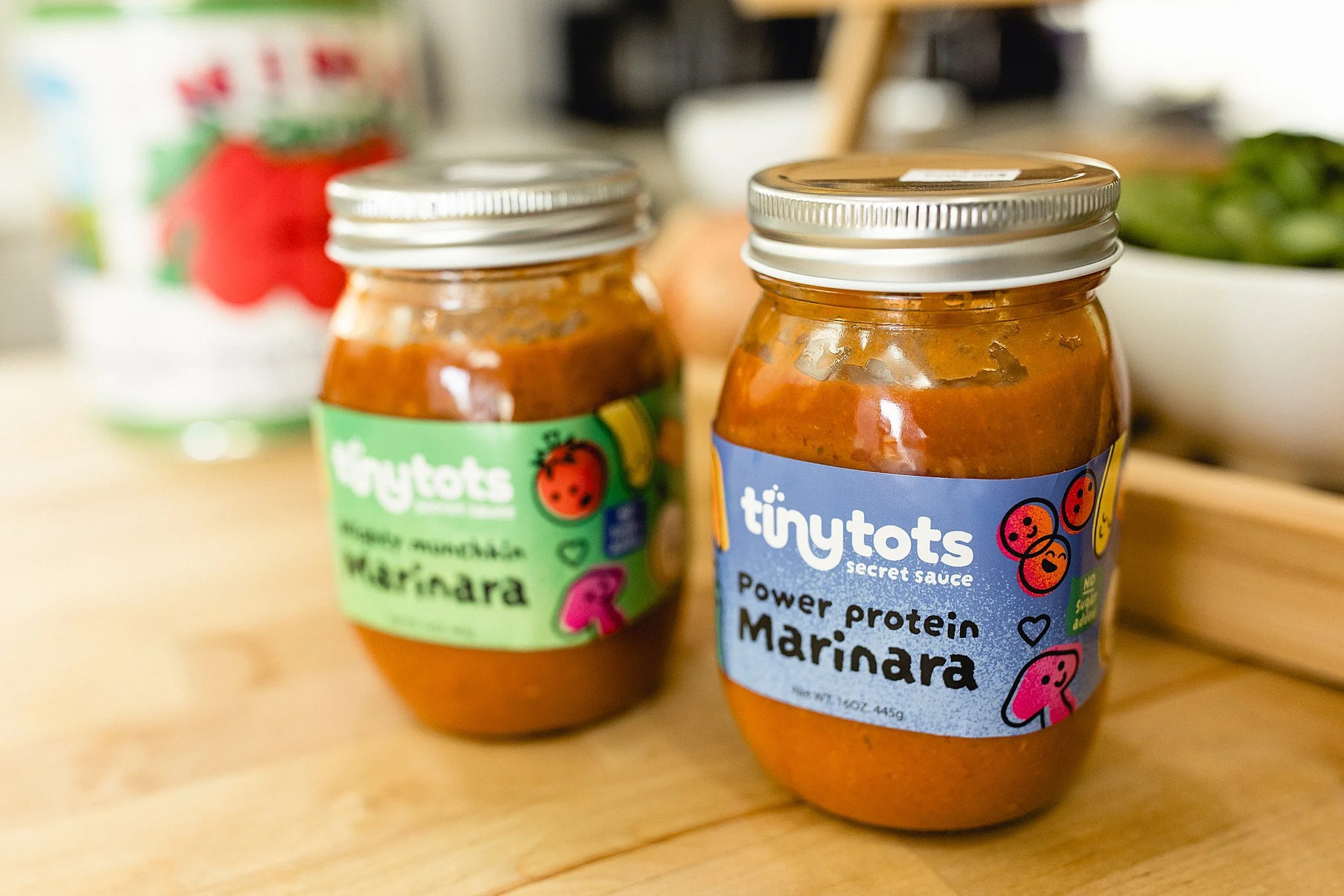Two jars of Tiny Tots secret sauce on a wooden surface, one labeled Power Protein Marinara, with cartoon tomato and mushroom illustrations, and a can of tomato puree in the background for a brand photoshoot with Ally and B Photography in Naperville.