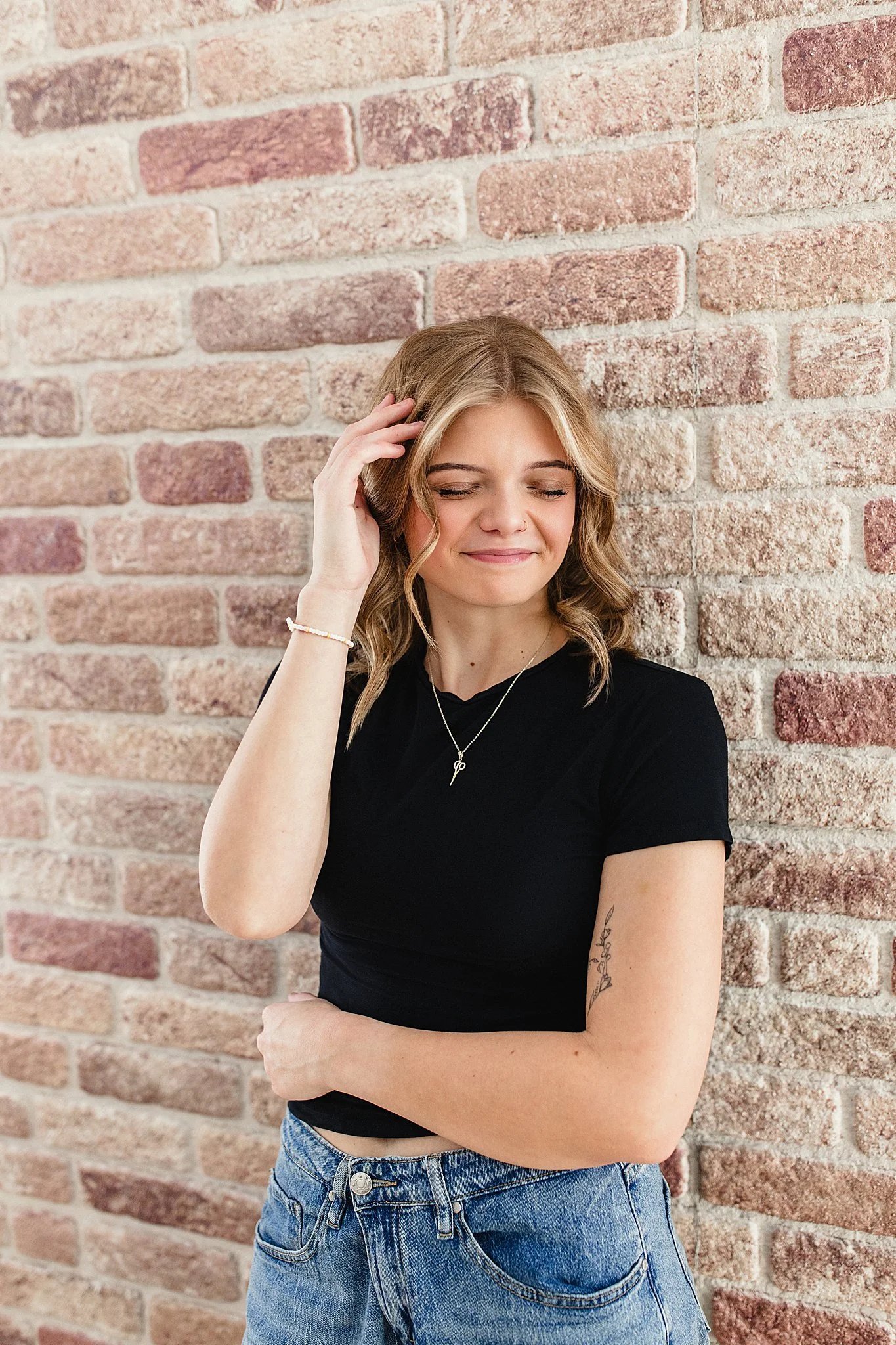 A young woman with shoulder-length wavy hair and fair skin stands against a red brick wall. She is smiling softly with her eyes closed, wearing a black t-shirt, blue jeans, a necklace, and a bracelet during a branding session in Naperville, IL.