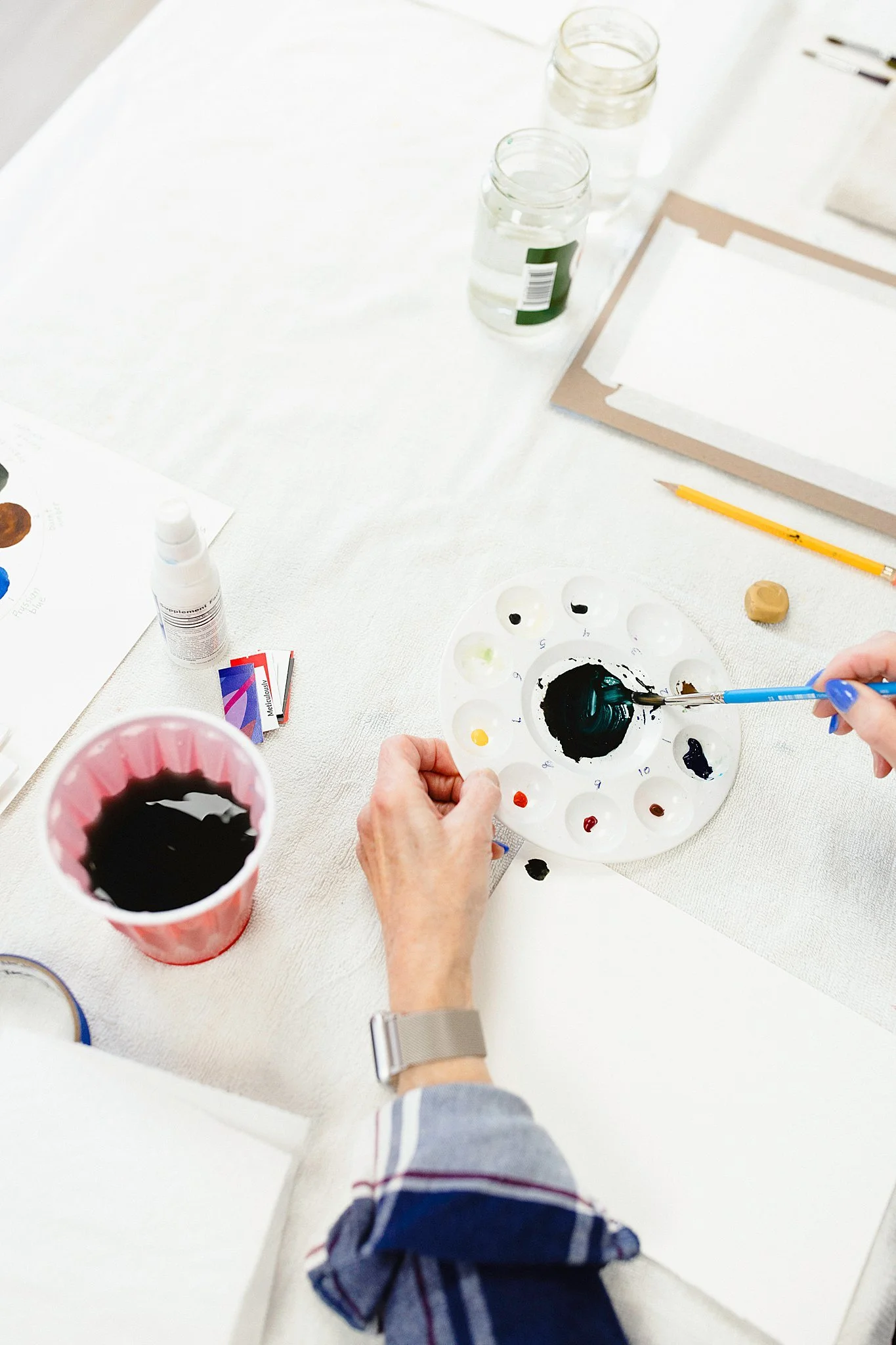 Someone mixing black paint on a circular palette during an art project, with various jars of water, a cup of black paint, and art supplies on a white table for artist branding photoshoot near me.