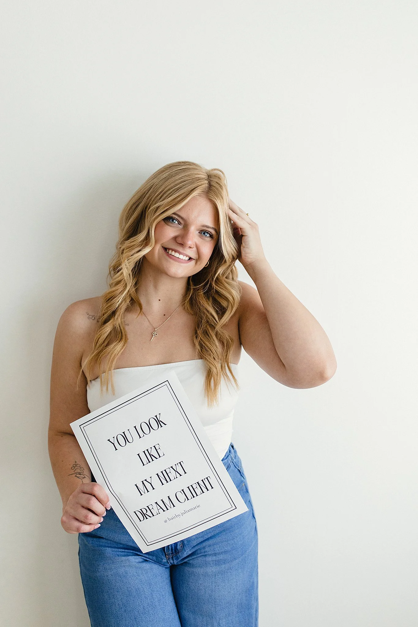 A smiling woman with long wavy blonde hair, wearing a white strapless top and blue jeans, holding a sign that says "You look like my next dream client" in front of a plain white wall with branding photographers in naperville il.