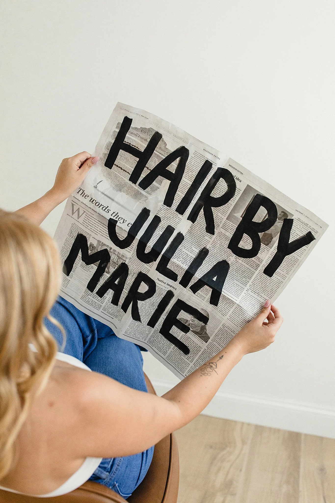 A person with long, curly blonde hair is sitting on a brown chair, holding a newspaper with large black letters painted on it that spell out "HAPPY BUM". The person is wearing blue jeans and a white top with brand photographers in Naperville.