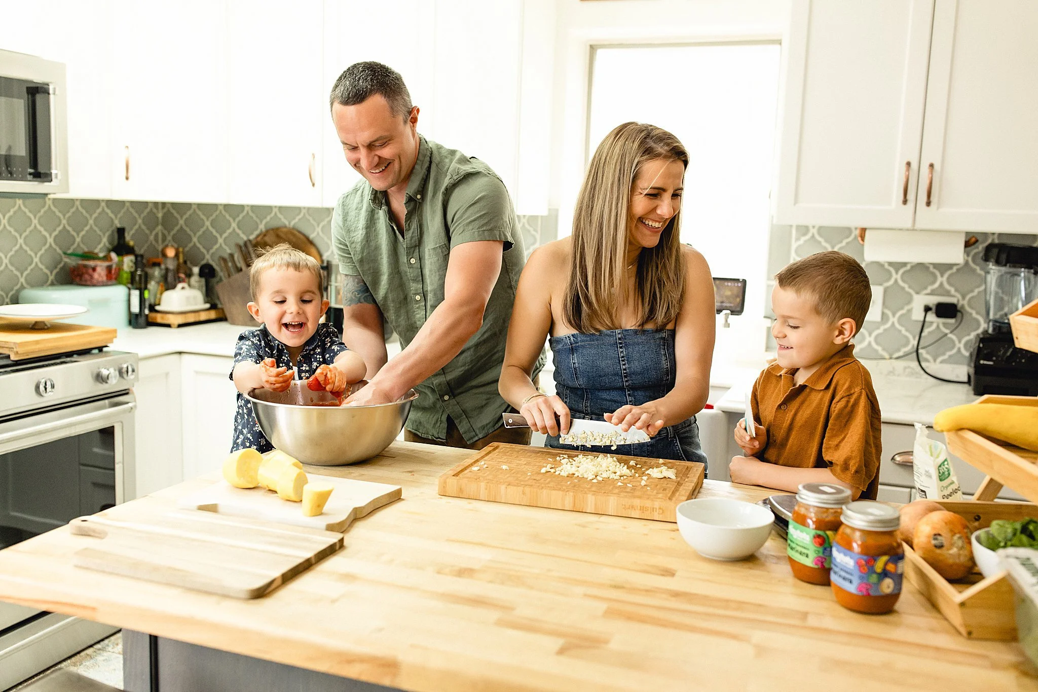 A family of four cooking together in the kitchen, with two children, a man, and a woman smiling and engaging in meal preparation during a small business branding session with Ally and B Photography.