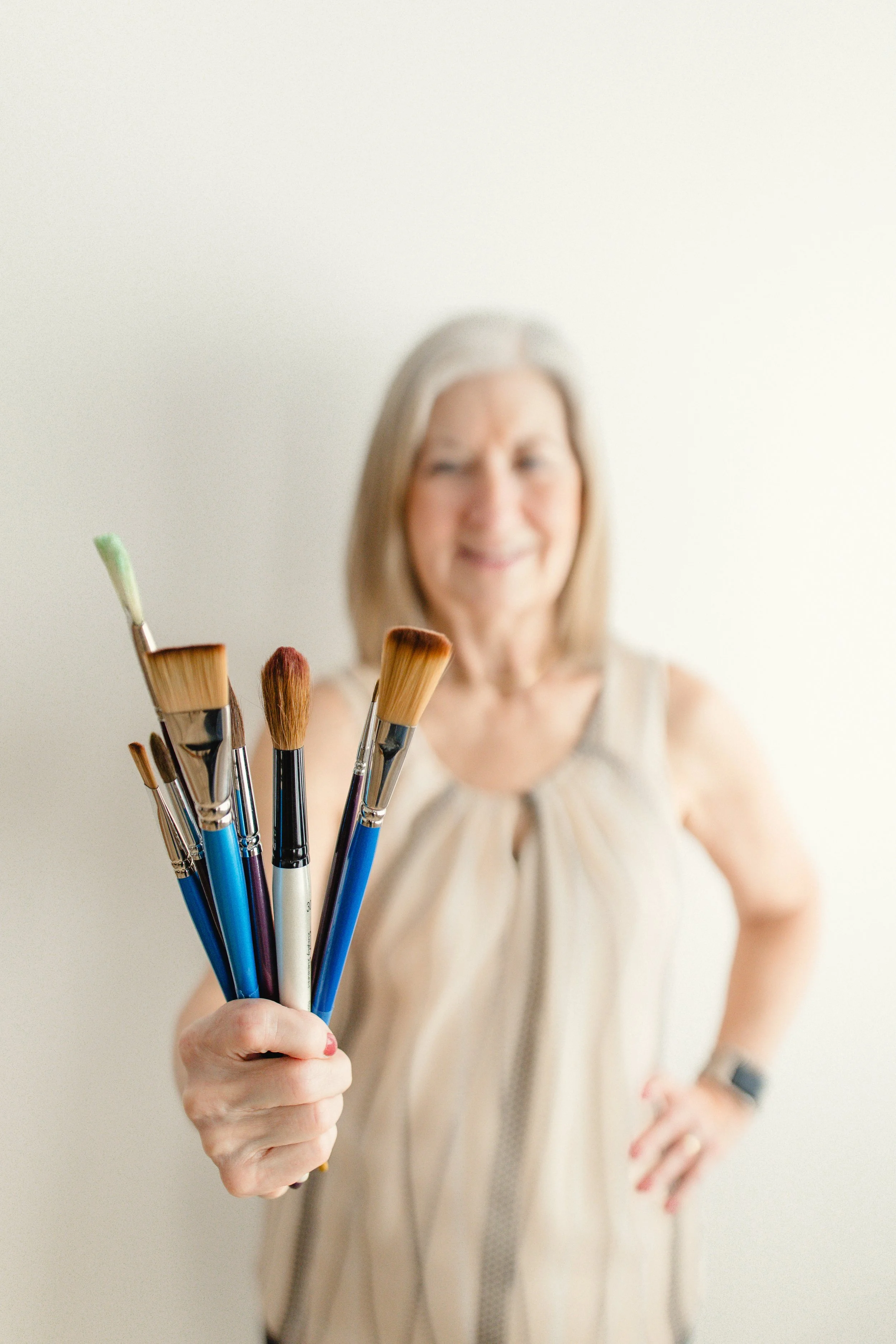 A woman holding a bunch of paintbrushes, with a blurry expression on her face in front of a plain white wall for branding photos Naperville.