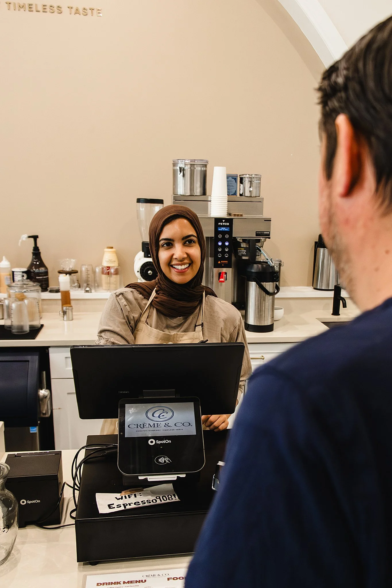 A woman wearing a brown hijab and beige apron smiling at a customer at a coffee shop counter. Behind her, there is coffee-making equipment and supplies on the counter. A man with short dark hair and a blue shirt is seen from the back in front of her.