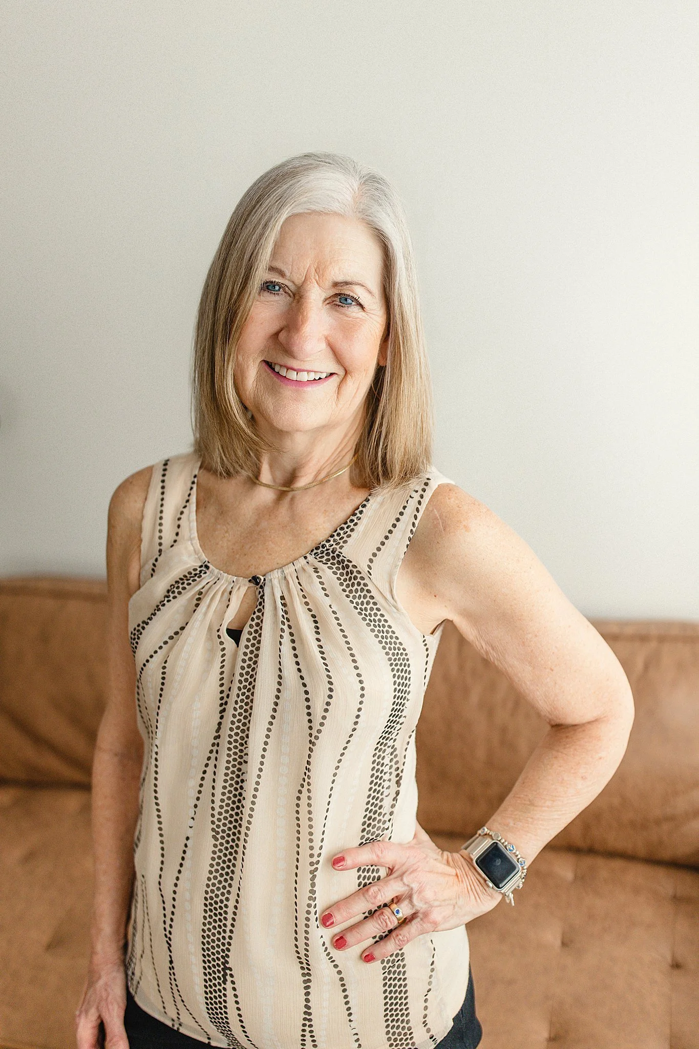 A smiling older woman with shoulder-length gray hair standing indoors. She wears a sleeveless beige top with black dotted patterns, a watch on her left wrist, and a ring on her right hand during headshots for a branding photoshoot in Naperville, IL.