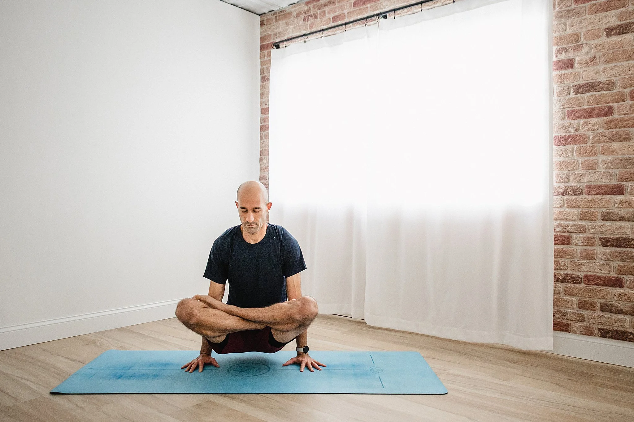 A man practicing yoga in a minimalist room with one white wall and one exposed brick wall, sitting on a yoga mat in a seated forward bend pose with legs crossed and hands on the ground for personal branding photos in Naperville, IL.