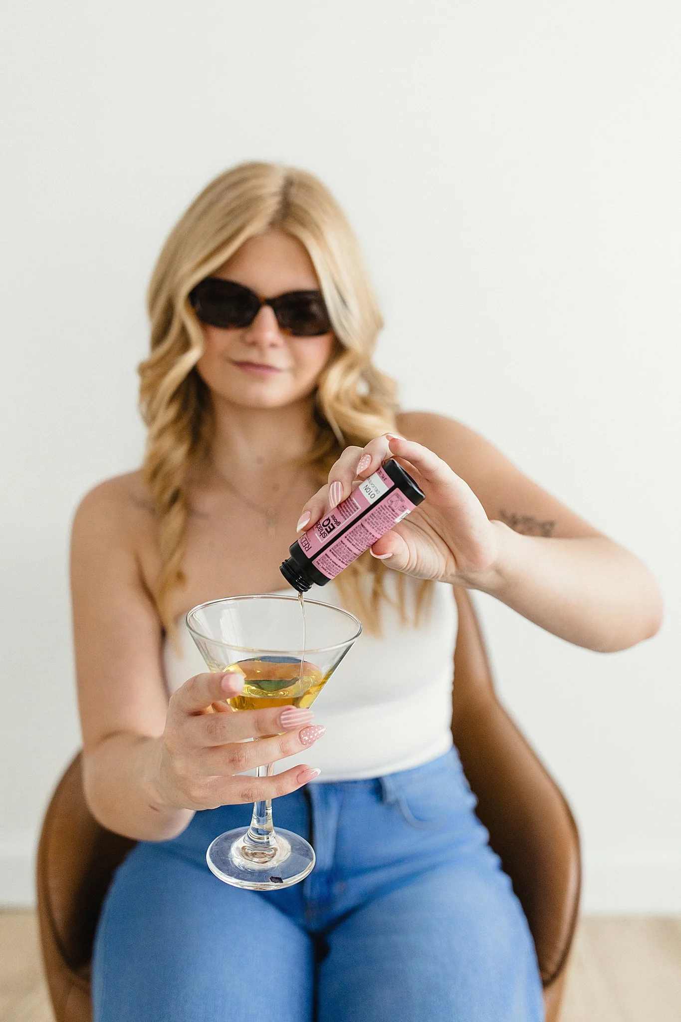A woman with blonde hair wearing black sunglasses and a white top, sitting on a chair, holding a martini glass with a yellow drink in one hand, and pouring a pink prescription pill bottle into the glass with the other hand for branding photos.