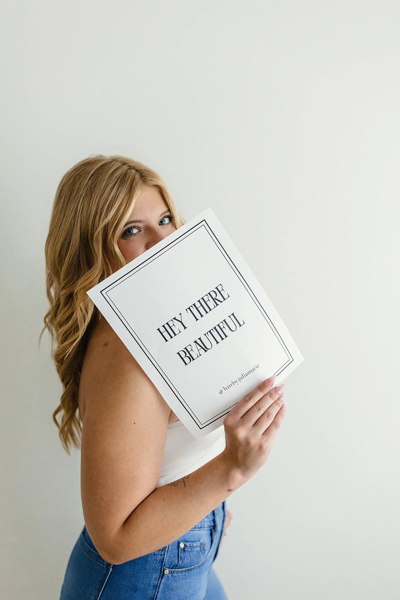 A woman with long blonde wavy hair holding a sign that says "Hey there, beautiful" in front of her face, wearing blue jeans and a strapless top, standing against a plain white wall during a branding photography session near me.