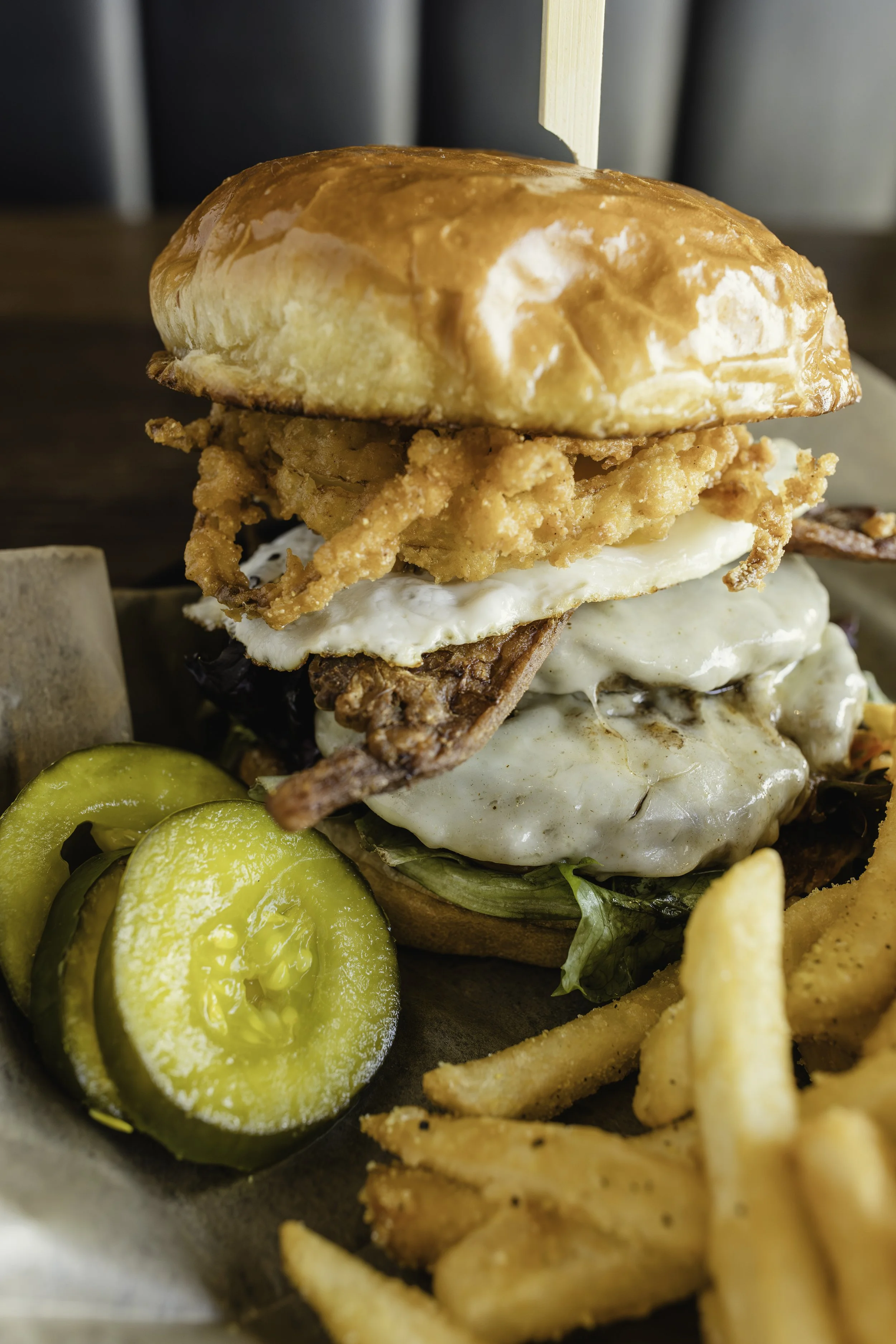 Close-up of a loaded burger with fried chicken, eggs, bacon, lettuce, and a brioche bun, served with pickles and fries for food photography in Naperville, IL.