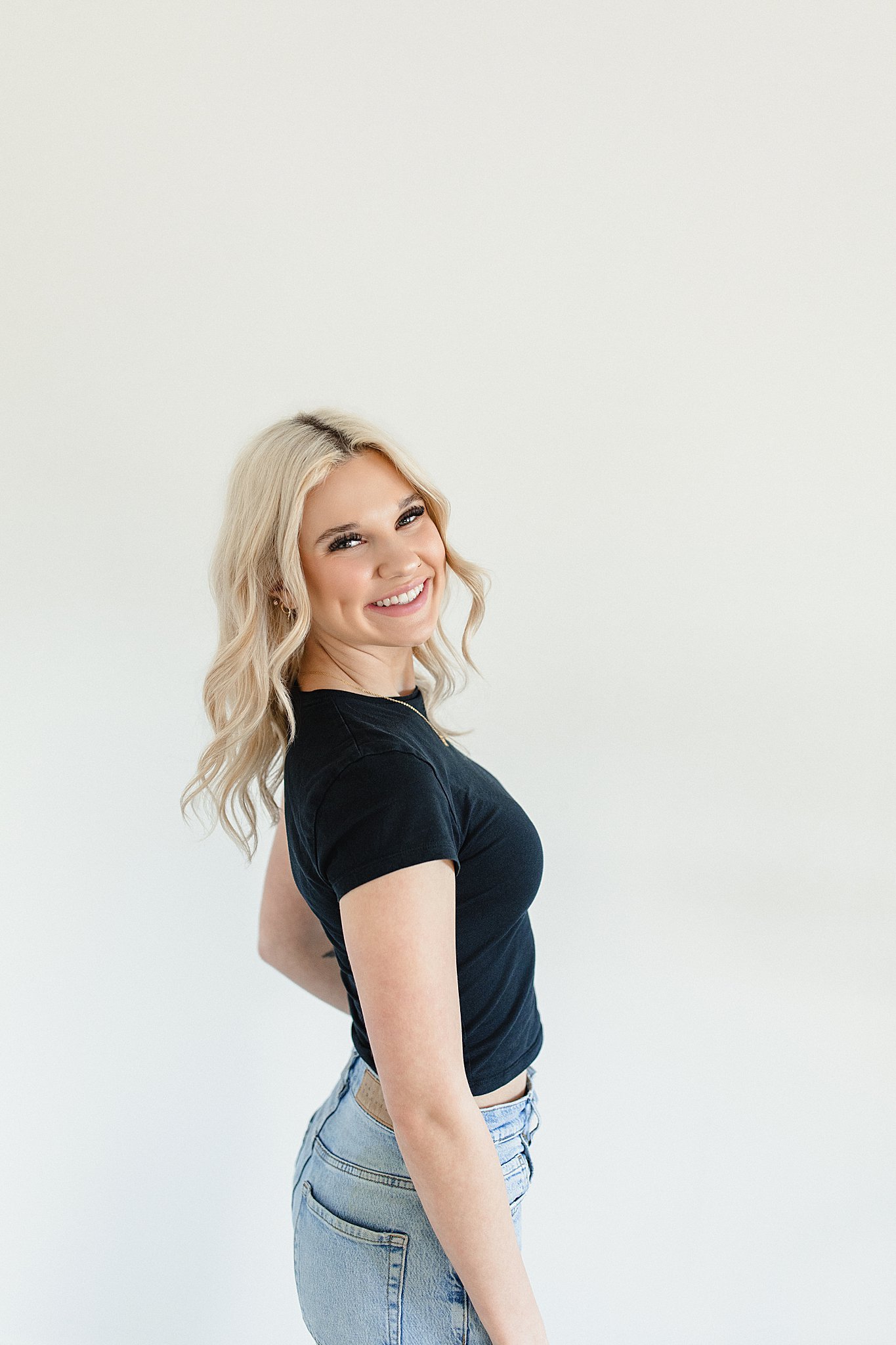 A young woman with blonde wavy hair smiling, wearing a black t-shirt and light blue jeans, standing against a plain white background with Naperville brand photographer Marie Lopez of Ally and B Photography.