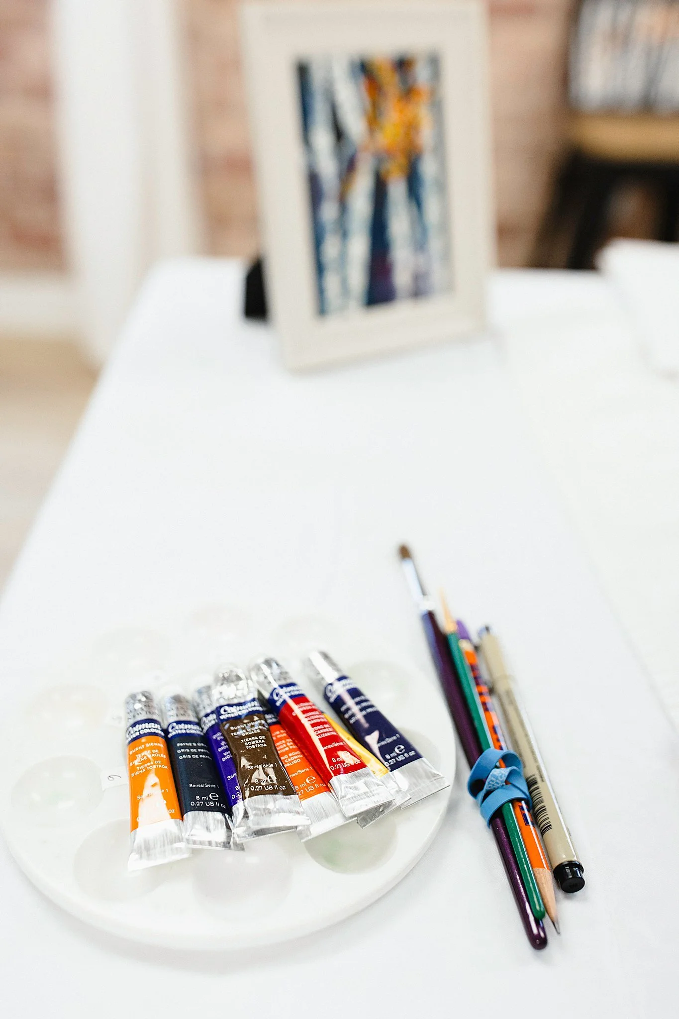 A palette of paint tubes, a pen, a mechanical pencil, and a paintbrush on a white table with a framed painting in the background for a watercolor paint flat lay photograph.