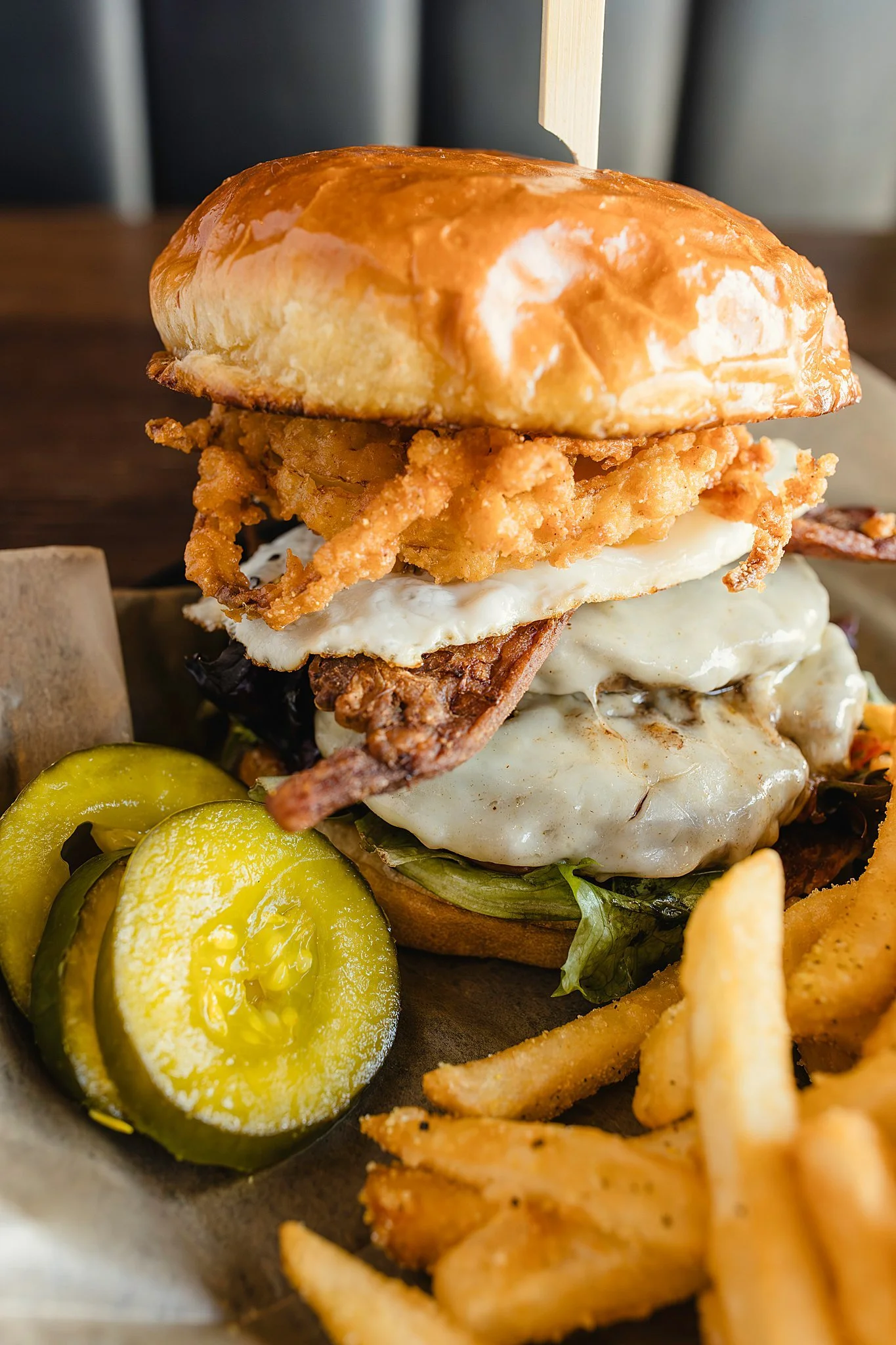 Close-up of a loaded burger with crispy fried onion rings, a fried egg, bacon, lettuce, and a toasted bun, served with pickles and French fries during food photography near me.