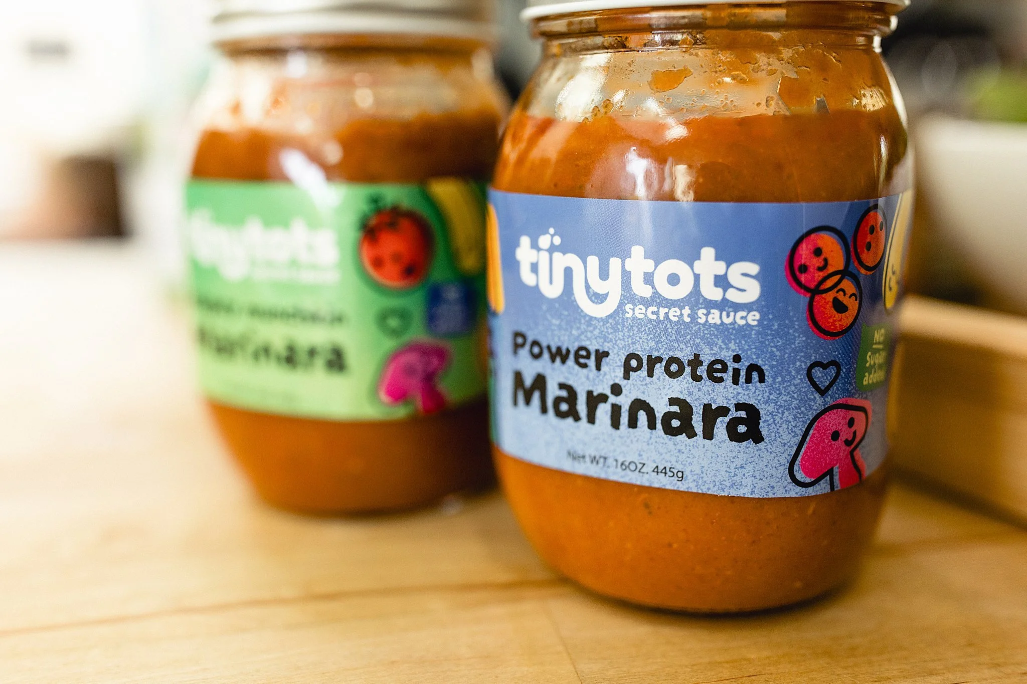 Two jars of tiny tots secret sauce, with one labeled Power protein Marinara and the other labeled Appetizer Marinara, placed on a wooden surface for Tiny Tots Secret Sauce product photography session with Ally and B Photography.