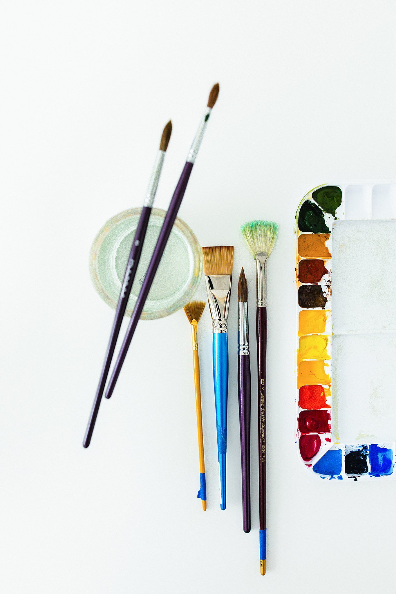 A set of watercolor painting supplies including paintbrushes, a palette with various colors, and a container of water on a white background for flat lay photographers in Naperville, IL.
