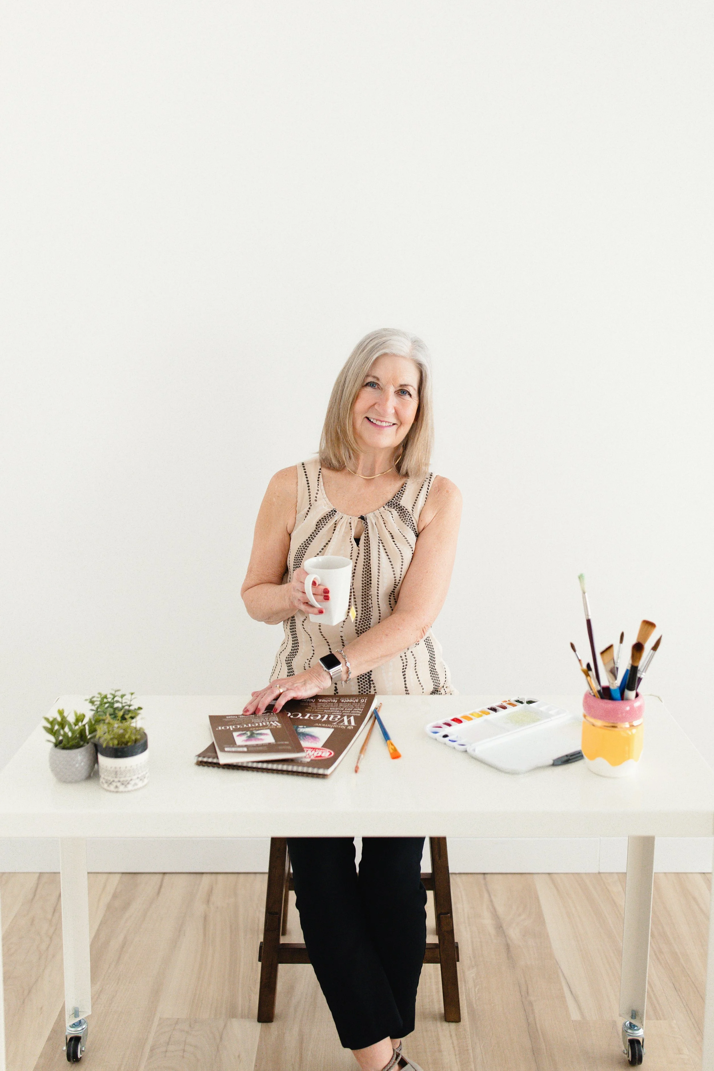 A smiling older woman with gray hair sitting at a white table, holding a white mug, surrounded by painting supplies, magazines, and potted plants for small business branding photography near me.