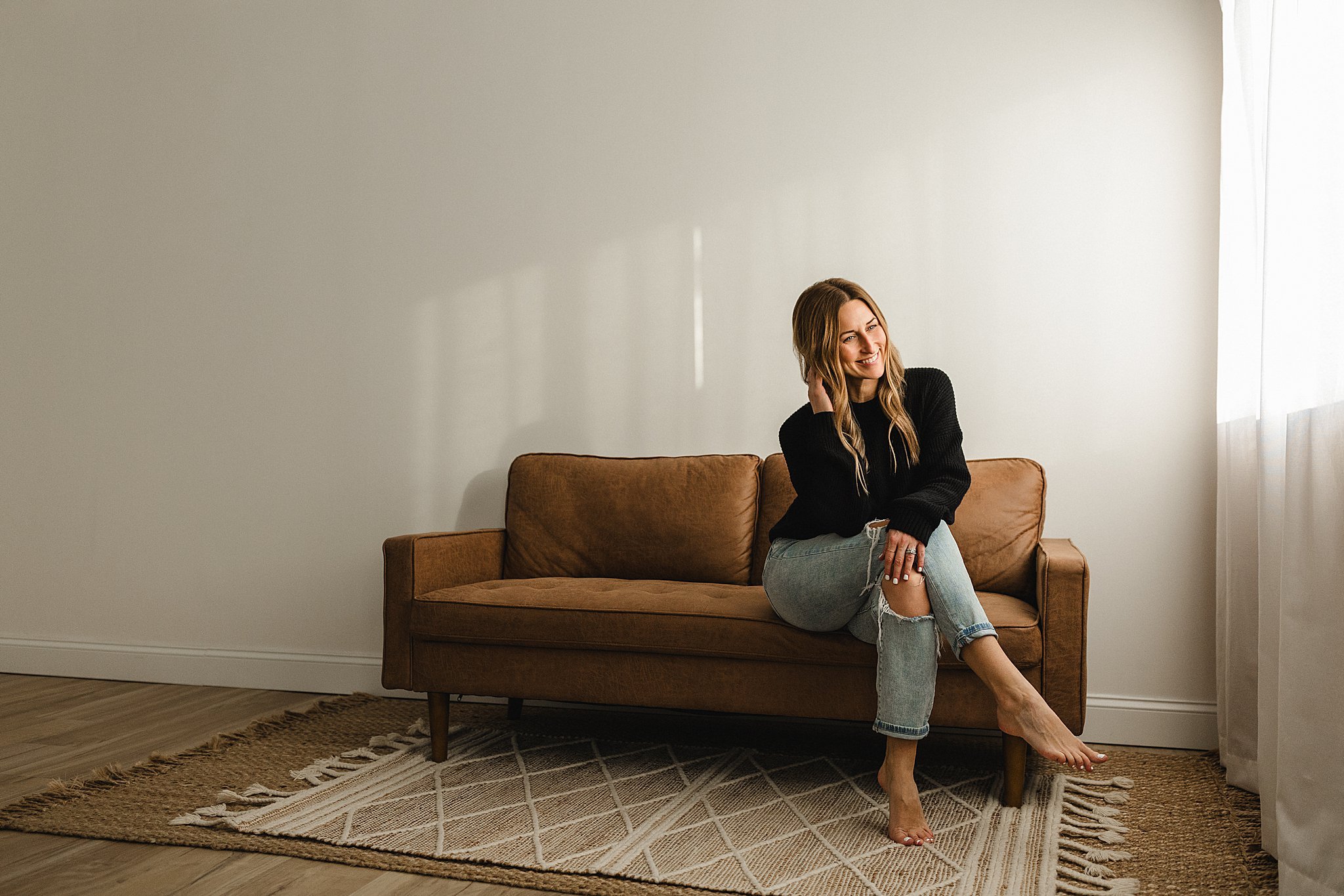 Marie Lopez of Ally and B Photography sitting on a brown couch in a minimalist room with white walls, blonde hair, wearing a black sweater and ripped jeans, smiling and looking to the side near a window with white curtains in her photography studio.
