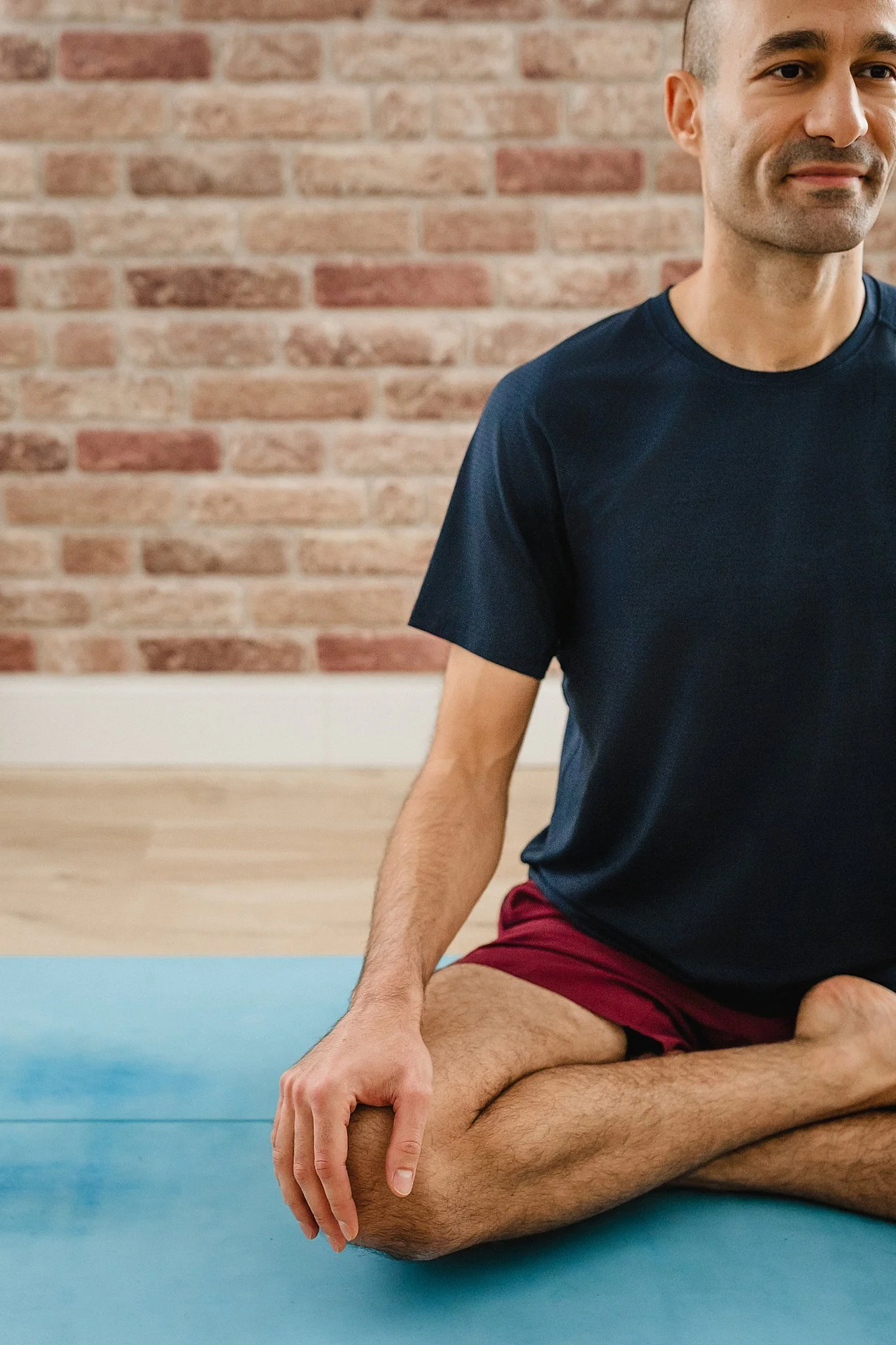 A man practicing yoga in a cross-legged seated position on a blue mat, with a red brick wall background during yoga brand photoshoot in Naperville, IL.