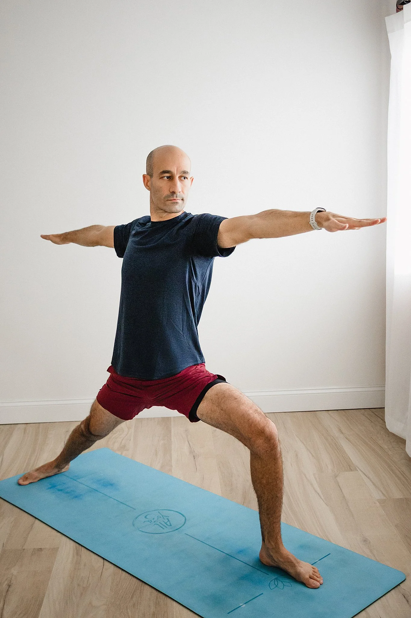 A man practicing yoga indoors on a blue yoga mat, standing in Warrior II pose with arms extended and legs wide apart during branding photos in Naperville, IL with Ally and B Photography.