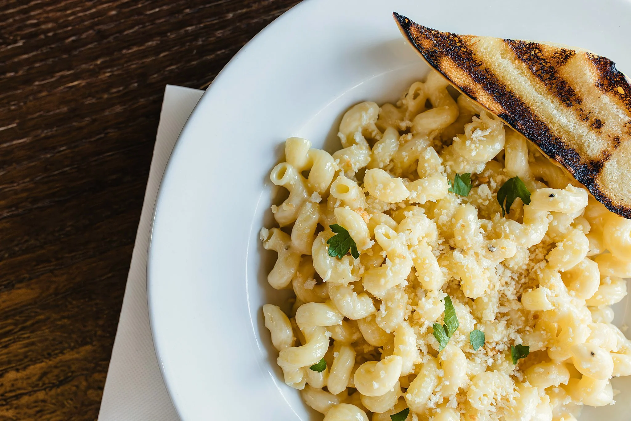 Creamy macaroni and cheese topped with grated cheese and fresh parsley, served with grilled bread on a white plate during a restaurant commercial photography session for food photography of menu items with Ally and B Photography.