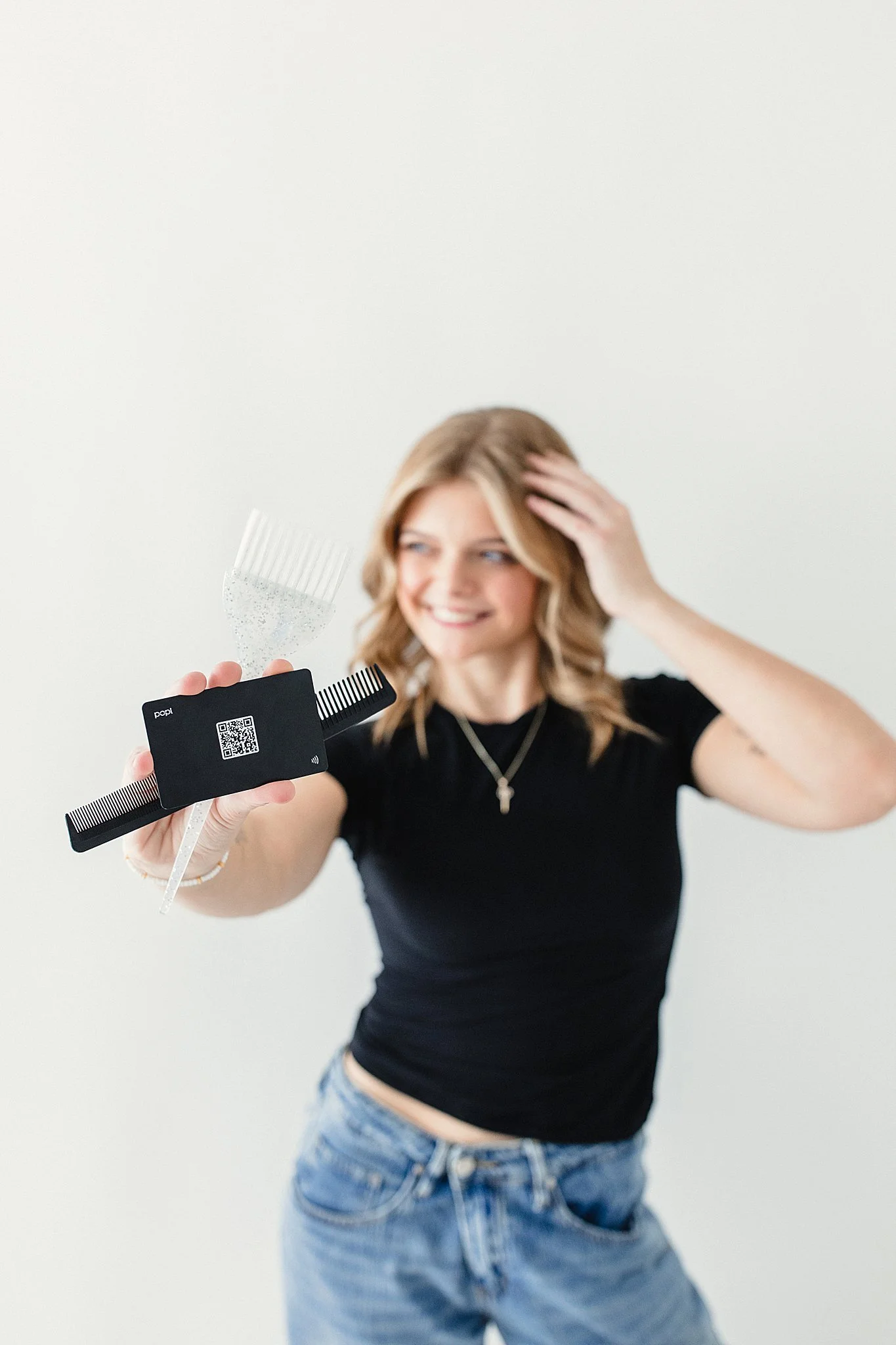 Young woman with blonde hair smiling, holding hair cutting tools including a comb, scissors, and a QR code tag, against a plain white background during a hairstylist branding session in Naperville, IL.