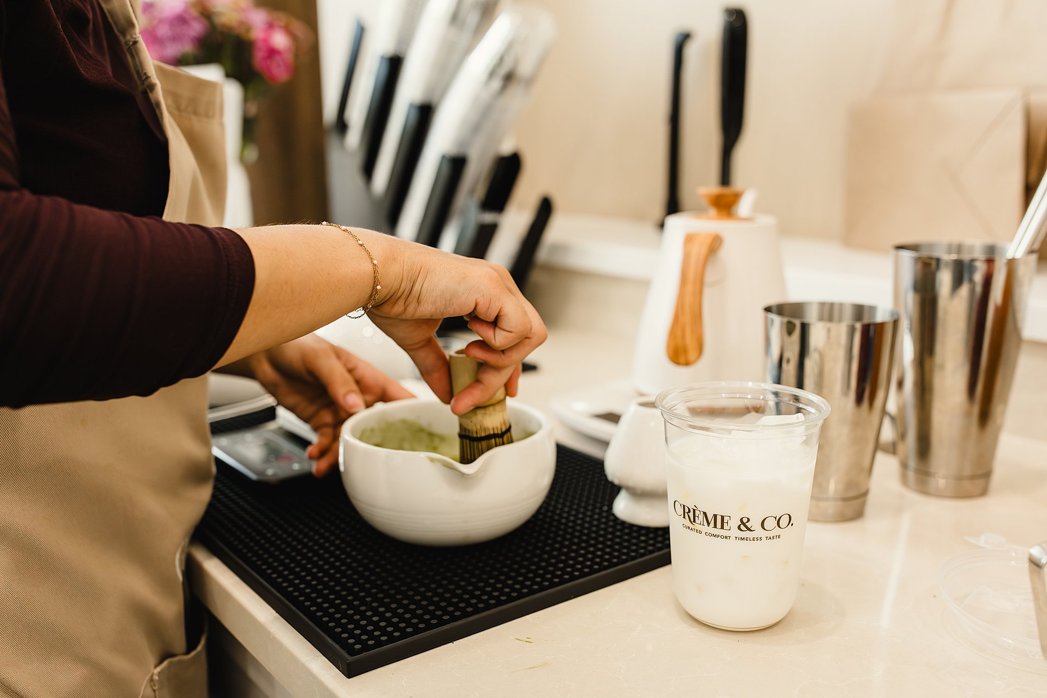 Person preparing food or drink at a kitchen counter with various utensils, a white container of a green paste, and a drink in a cup labeled 'CREME & CO.' for coffee shop branding photos.