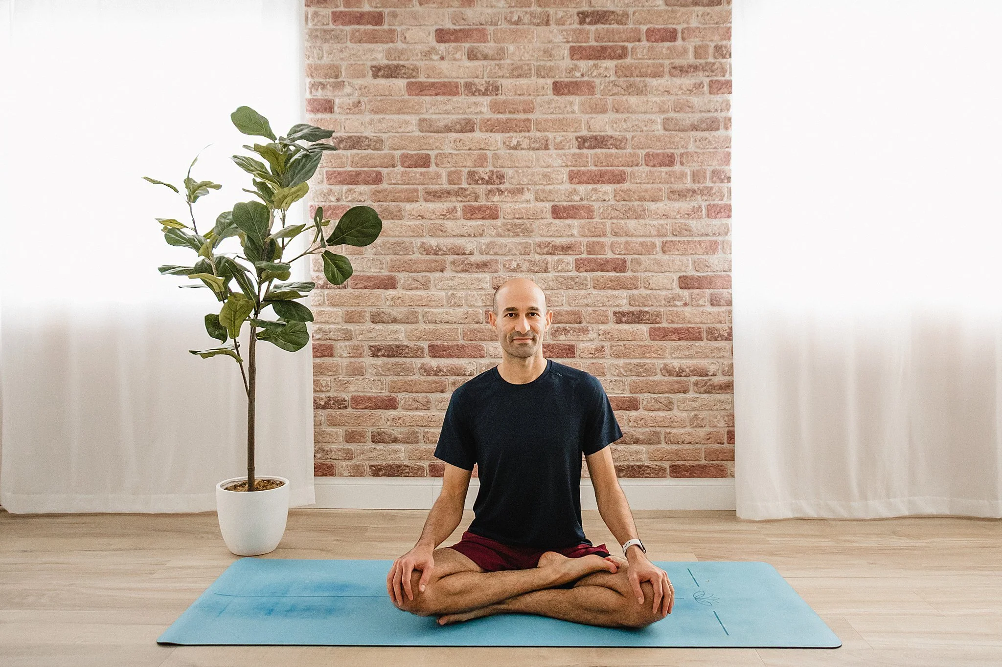 A man sitting cross-legged on a yoga mat, meditating in front of a brick wall inside a room with white curtains and a potted plant with Naperville brand photographer.