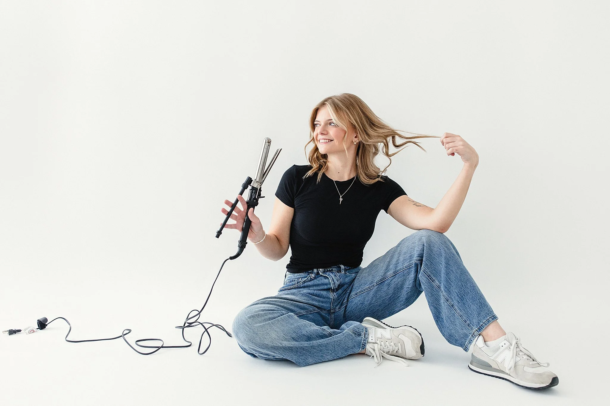 A young woman sitting on the floor, holding a curling iron, smiling, with hair styling tools and wires around her, against a plain white background for brand photos by Ally and B Photography in Naperville, IL.
