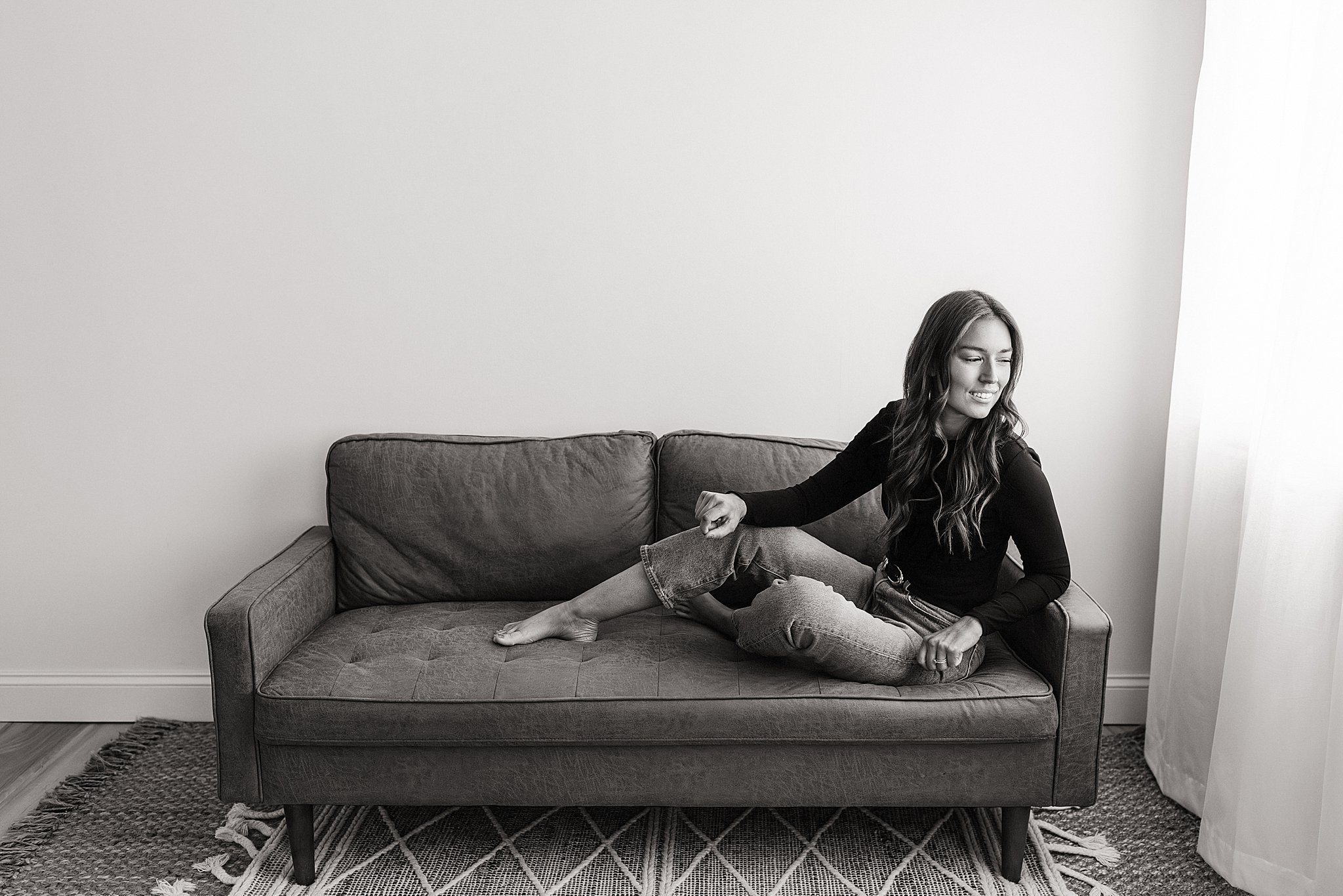 A young woman with long wavy hair sitting on a sofa, smiling, with her right leg crossed over her left, in front of a plain white wall and curtains on the right during a personal branding session with Ally and B Photography in Naperville, IL.