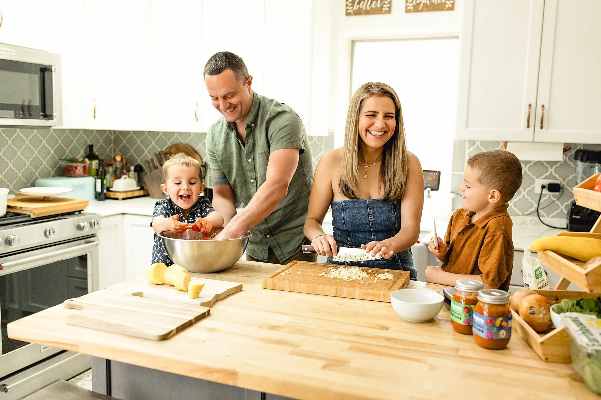 A happy family in the kitchen preparing food together. A man and a young girl are mixing ingredients in a bowl, while a woman and a boy are chopping vegetables on a cutting board, all smiling during a branding photoshoot with Ally and B Photography.