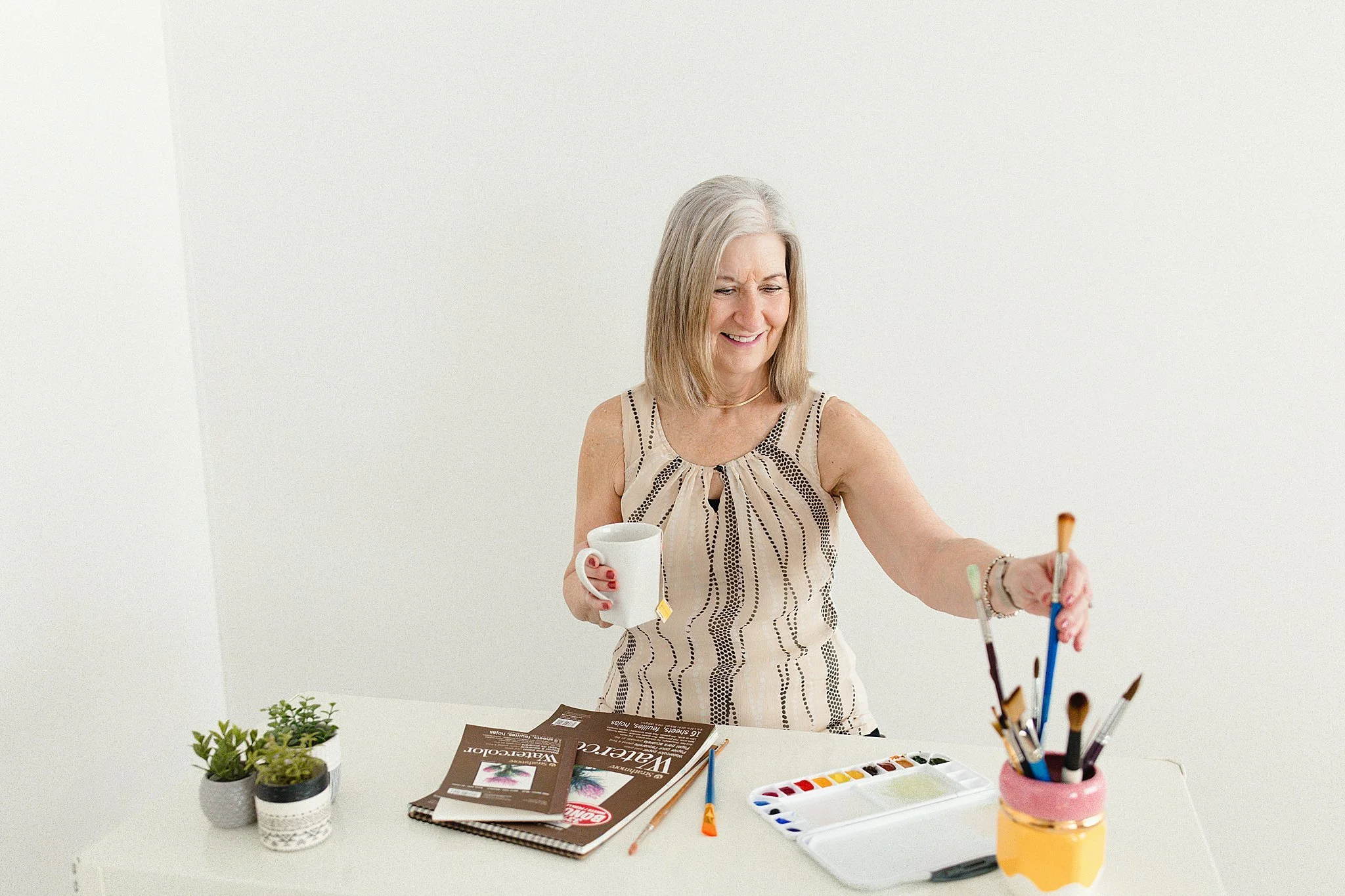 A smiling middle-aged woman with gray hair, wearing a sleeveless beige and black patterned top, is holding a white mug with a tea bag tag and a paintbrush and watercolor paints while standing at a white table for brand photography in Naperville, IL.