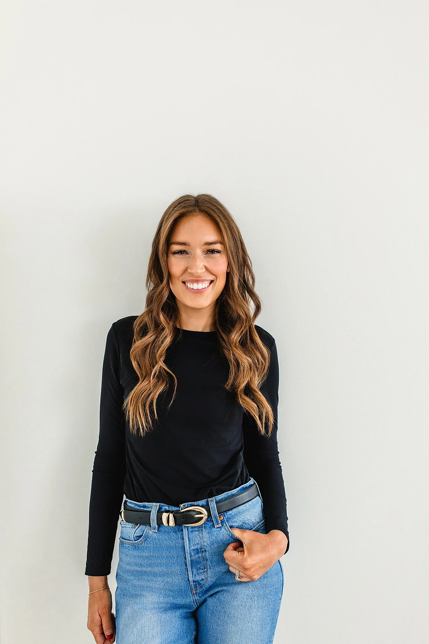A young woman with long, wavy brown hair, smiling and wearing a black long-sleeve shirt, light blue jeans, and a black belt with a gold buckle, standing against a plain white wall during a personal branding session in Naperville, IL.
