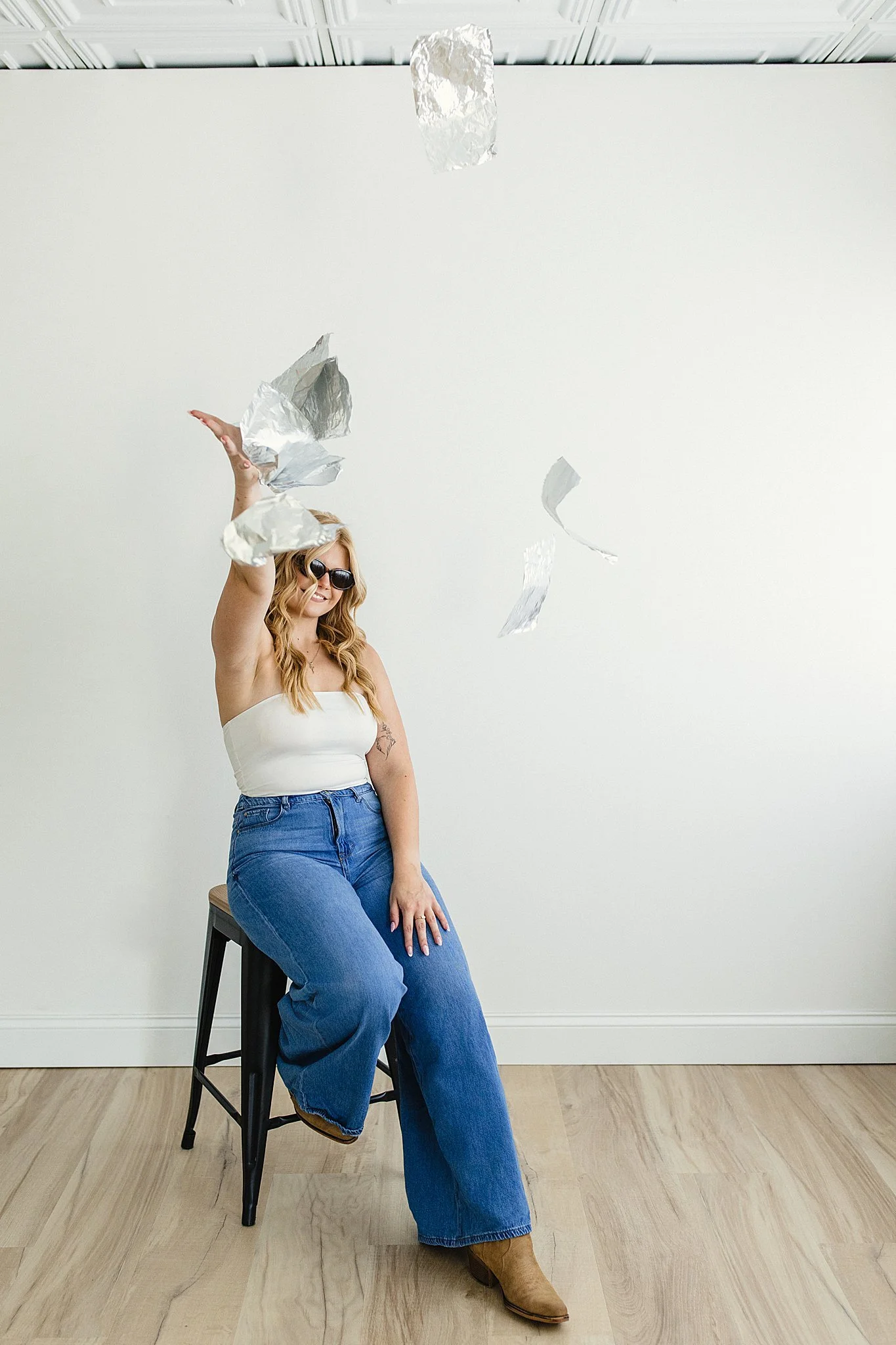 A woman with long wavy red hair and sunglasses sitting on a black stool, wearing a white strapless top, blue jeans, and brown boots. She is inside a room with light wooden flooring and a white wall background. Several pieces of crumpled aluminum foil