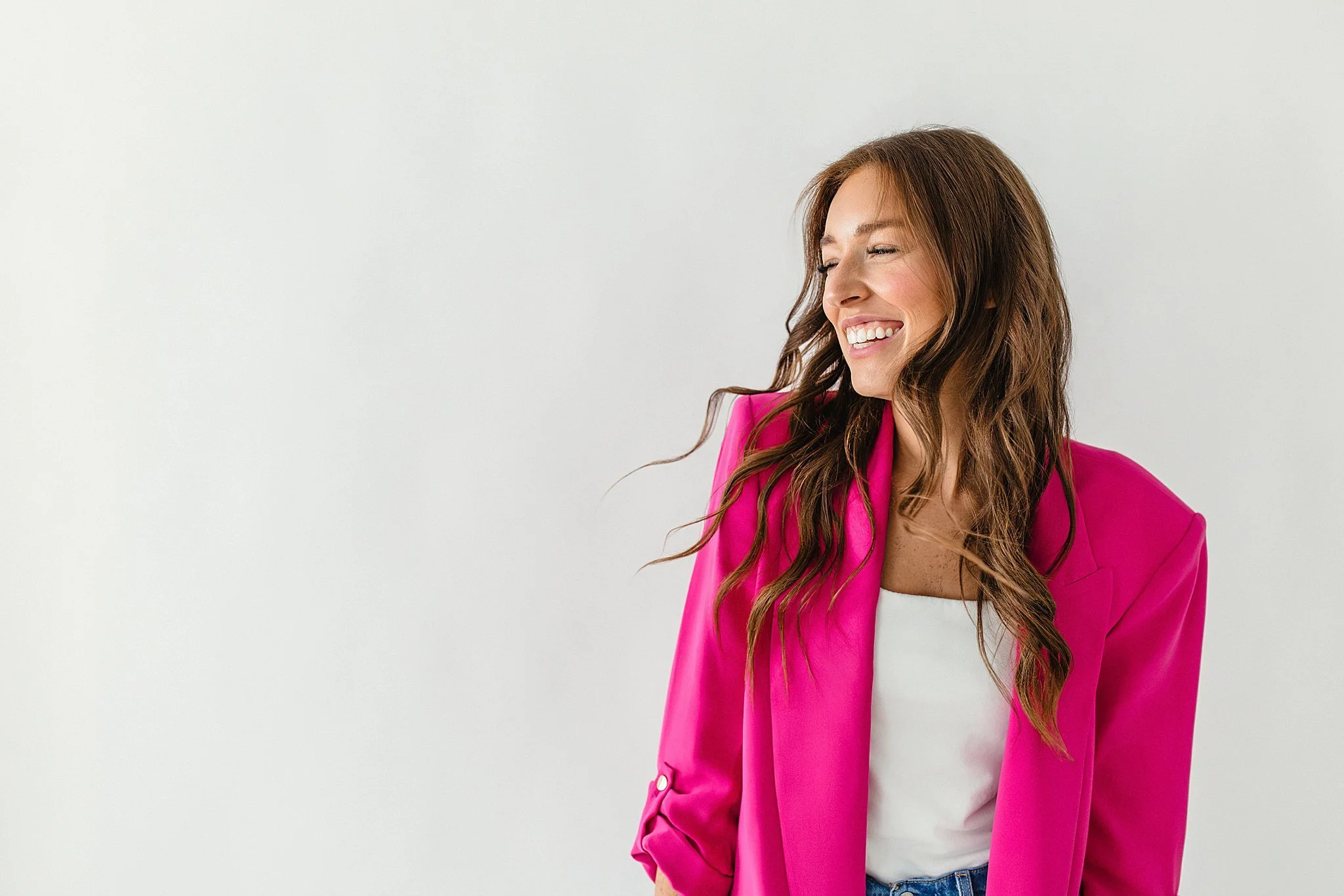 Smiling woman with long wavy brown hair wearing a bright pink blazer and white top, standing against a plain white background during a Naperville brand photoshoot.