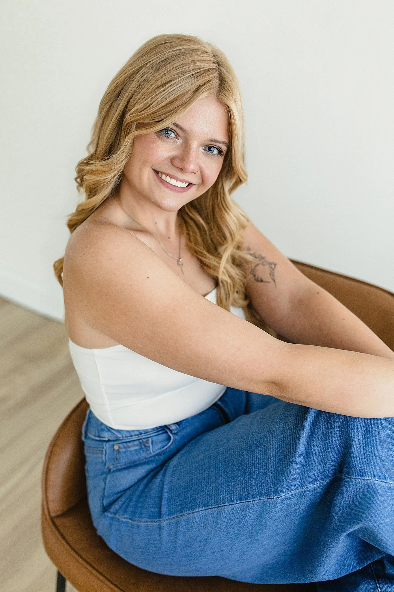 A woman with blonde hair and blue eyes sitting on a brown chair, smiling, wearing a white tank top and blue jeans, with a tattoo on her upper arm during a personal branding session in downtown Naperville with Ally and B Photography.