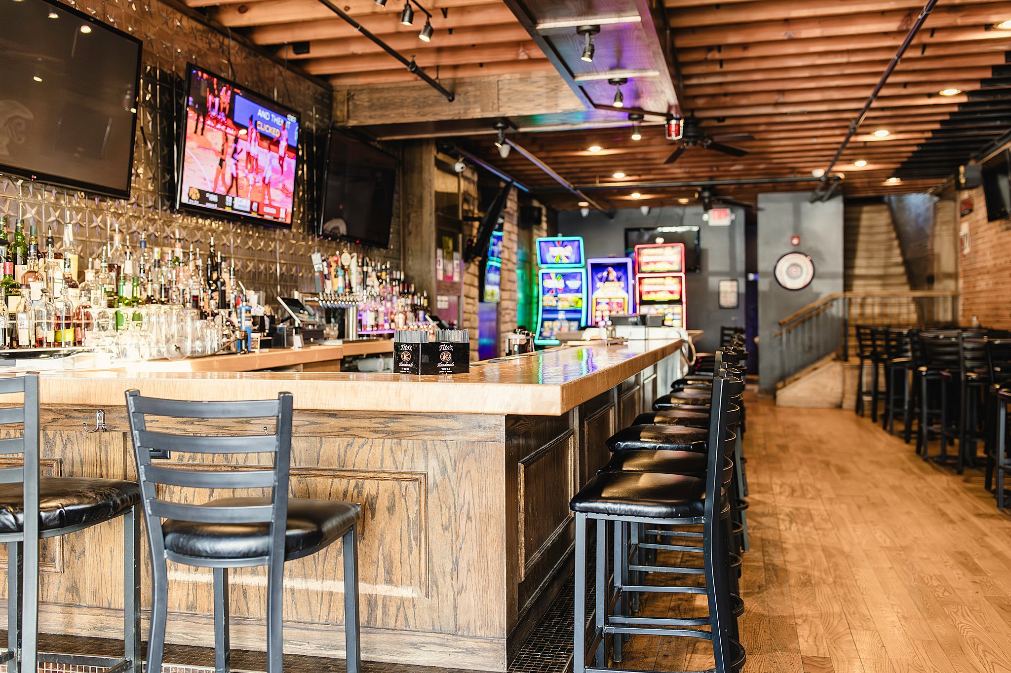 Empty bar with high black leather stools along a wooden counter in a dimly lit pub with TV screens and slot machines in the background during a bar branding photography session with Ally and B Photography brand photography.