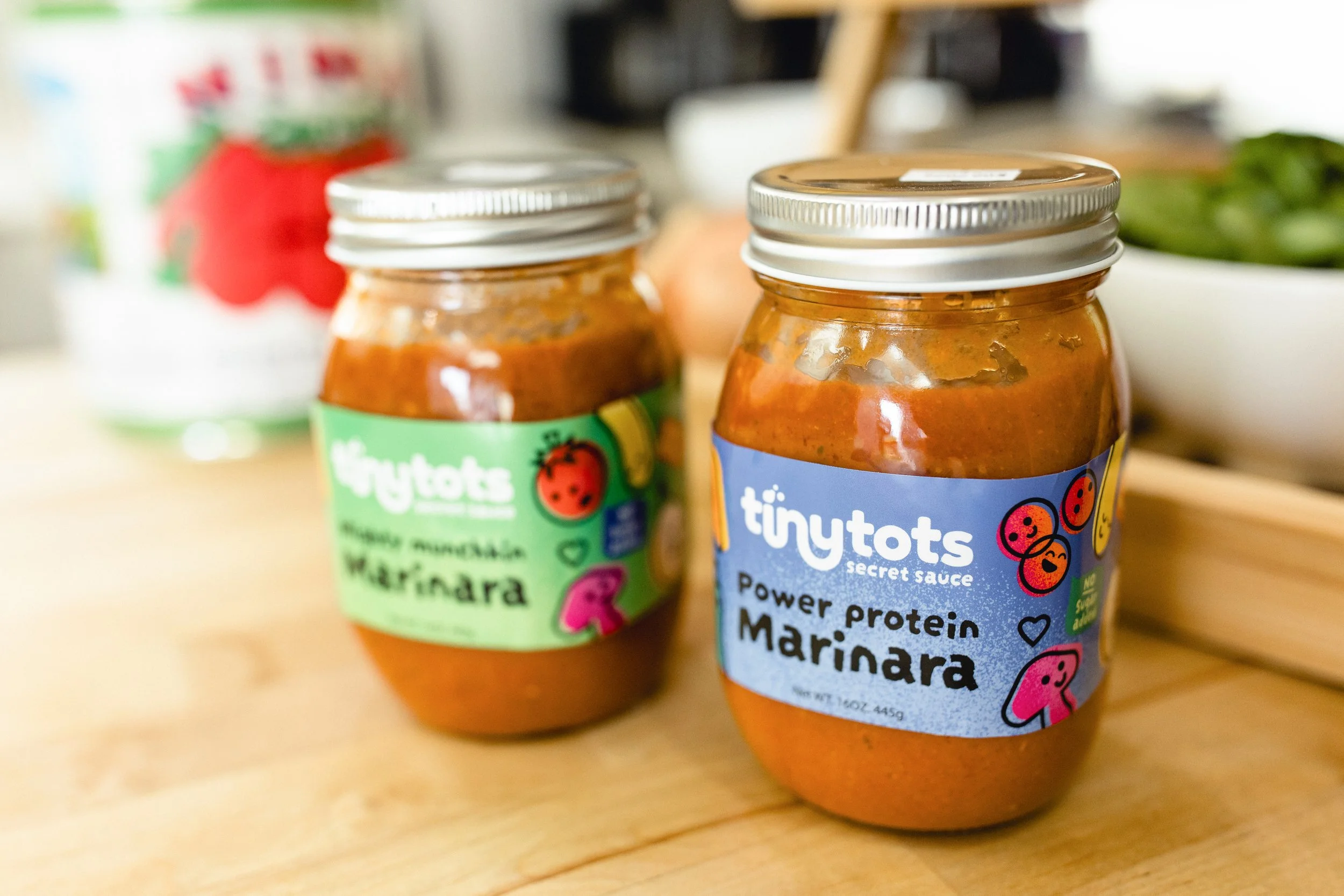 Two jars of tiny tots marinara sauce, one labeled 'Waterdr,' the other 'Marinara,' on a wooden surface for product photography near me.