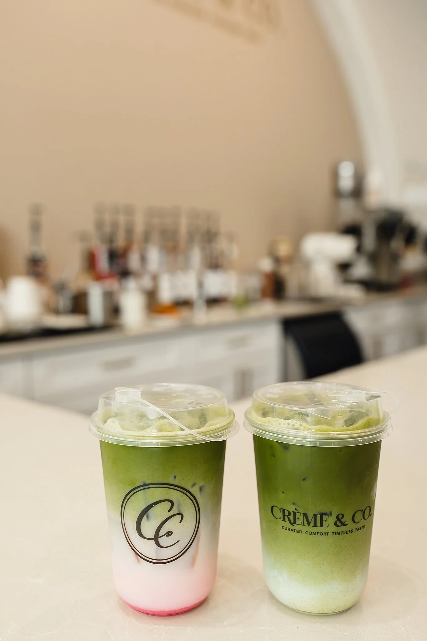 Two cold beverages in green cups with clear plastic lids and straws, one light pink and the other light green, on a white surface, with a blurry background of a bar or counter with bottles and equipment with food and drink photographers in Naperville
