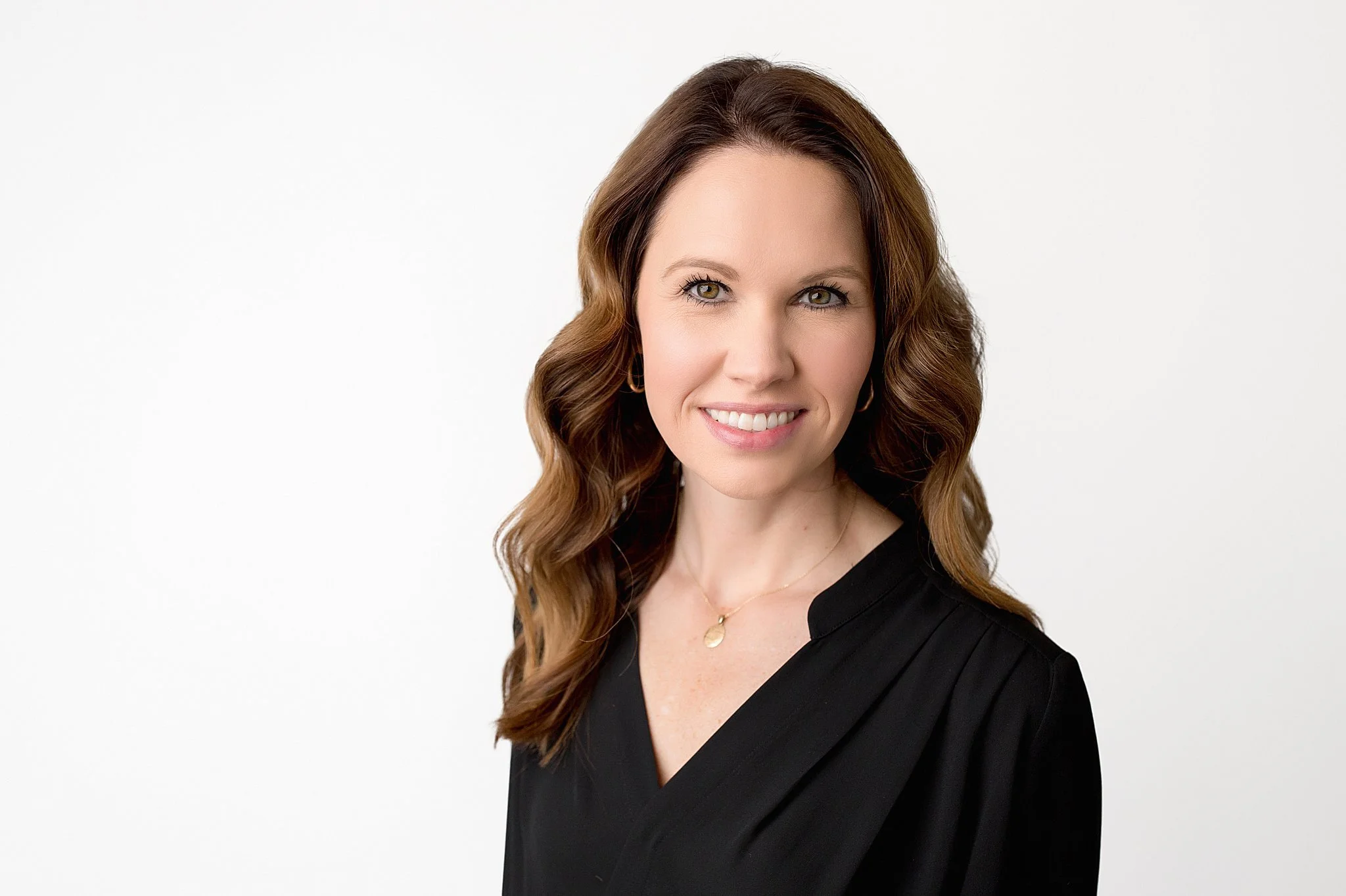 Portrait of a woman with wavy brown hair, wearing a black top and a gold necklace, smiling against a plain white background for best headshot photographers Naperville.