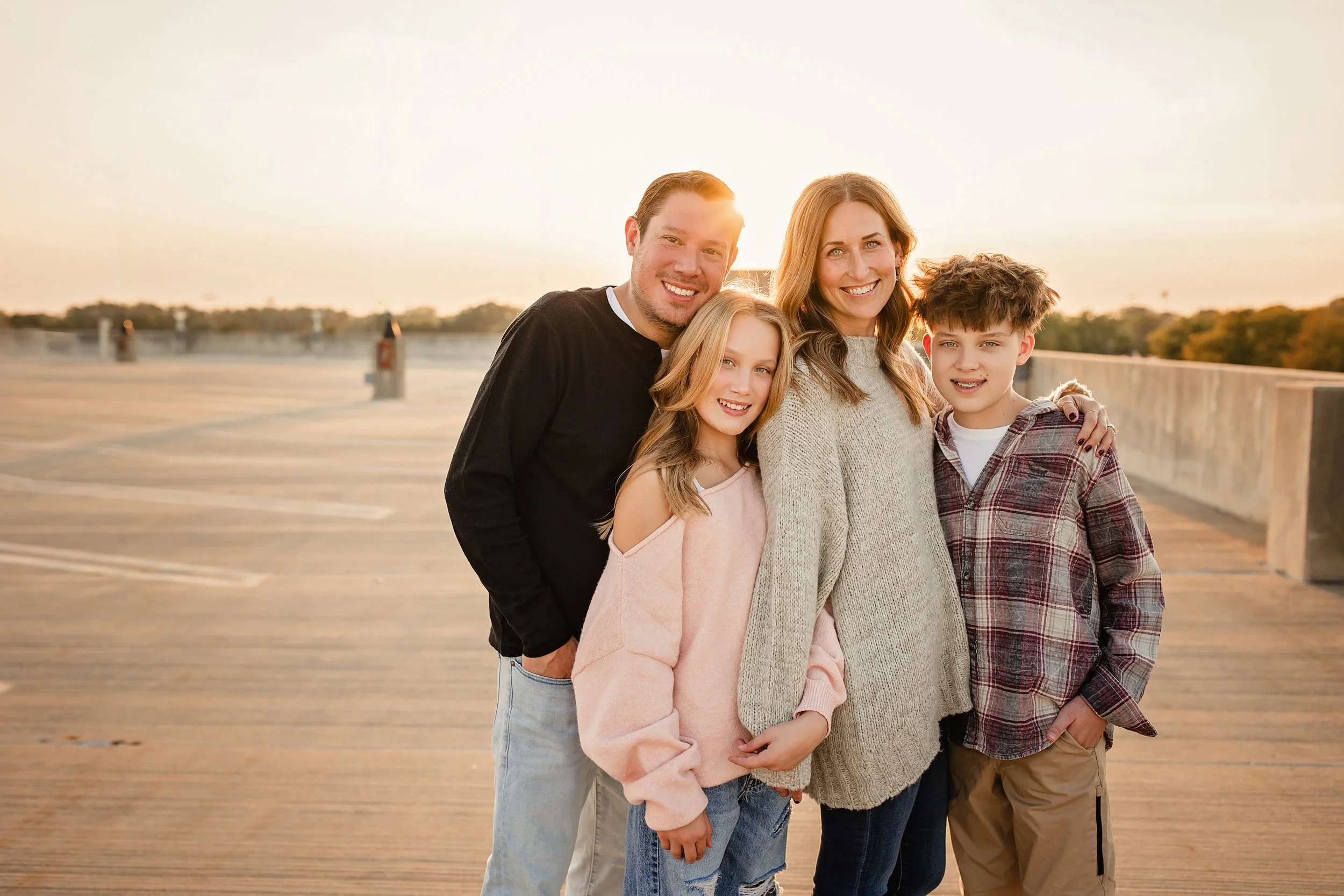 Family of four standing on a wooden parking garage rooftop at sunset, smiling during a Naperville family photography session of Marie Lopez of Ally and B Photography and her family