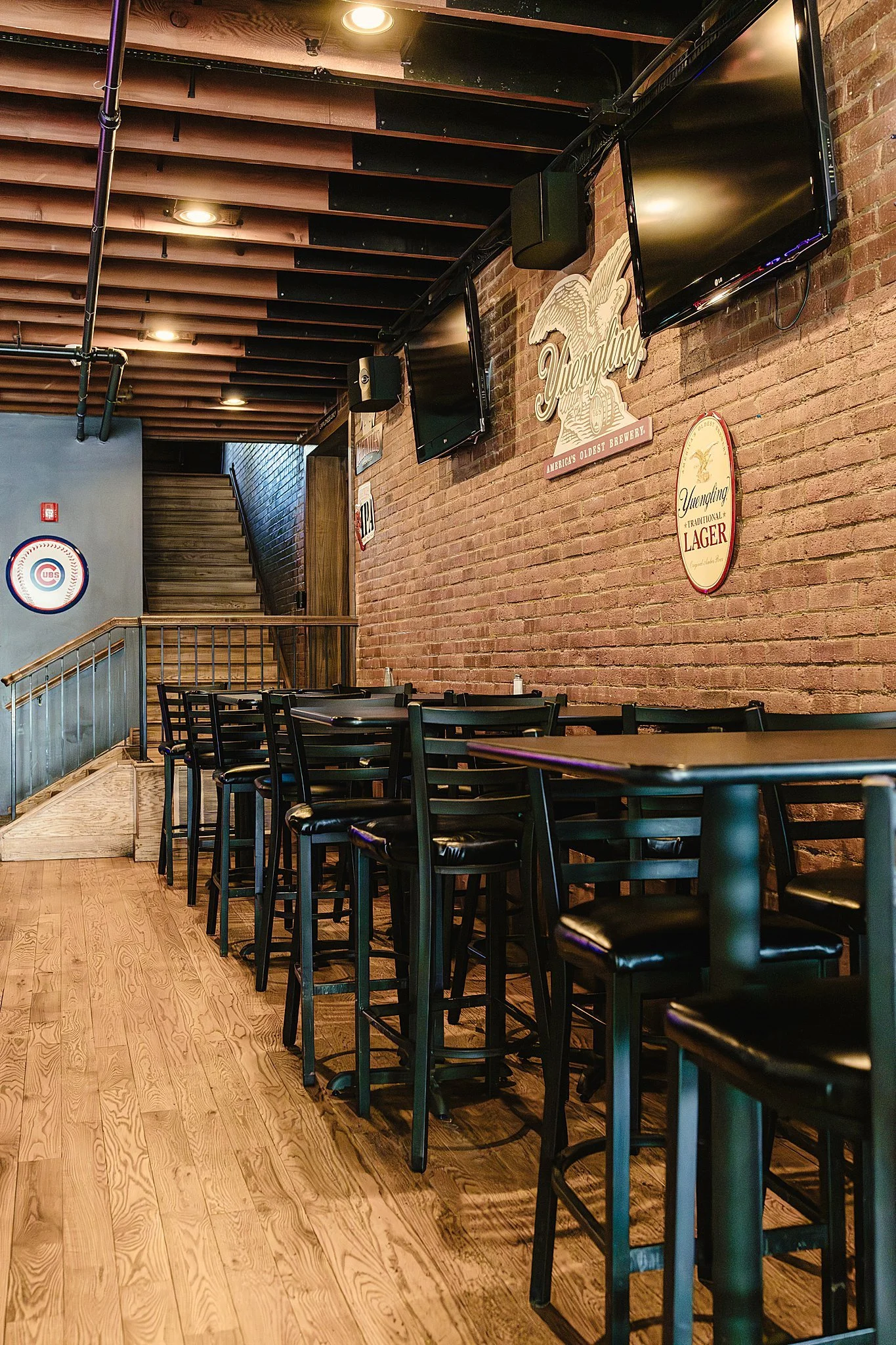Interior of a brewery or bar with wooden flooring, brick walls, black chairs and tables, two TV screens on the wall, and brewery signs for bar branding photos near me.