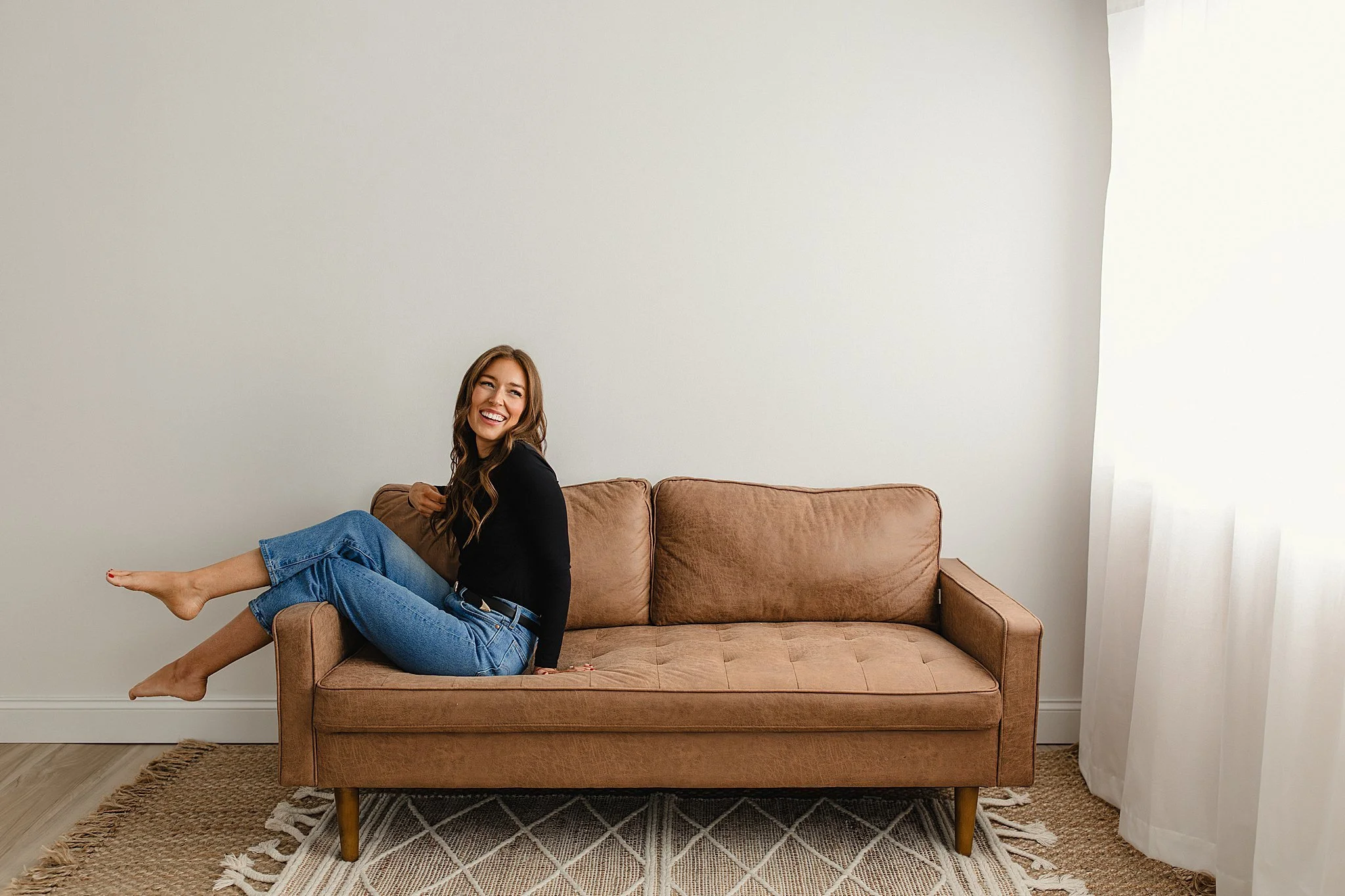 A woman with long brown hair smiling and sitting sideways on a brown sofa, wearing a black top and blue jeans, in a room with a white wall, a beige rug, and a large window with white curtains during a personal branding session in Naperville, IL.