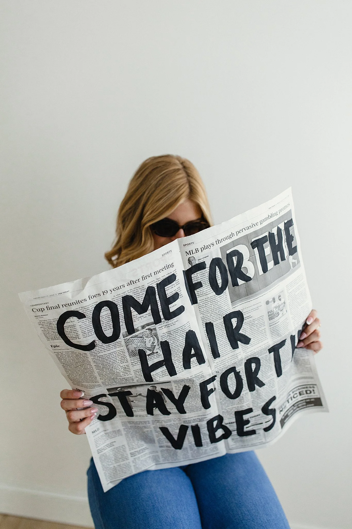 A woman with blonde hair wearing sunglasses and blue jeans sitting on the floor against a plain white wall, holding a newspaper that covers her face, with large black text on the front page saying 'COME FOR THE HAIR' 'STAY FOR THE VIBES.' 