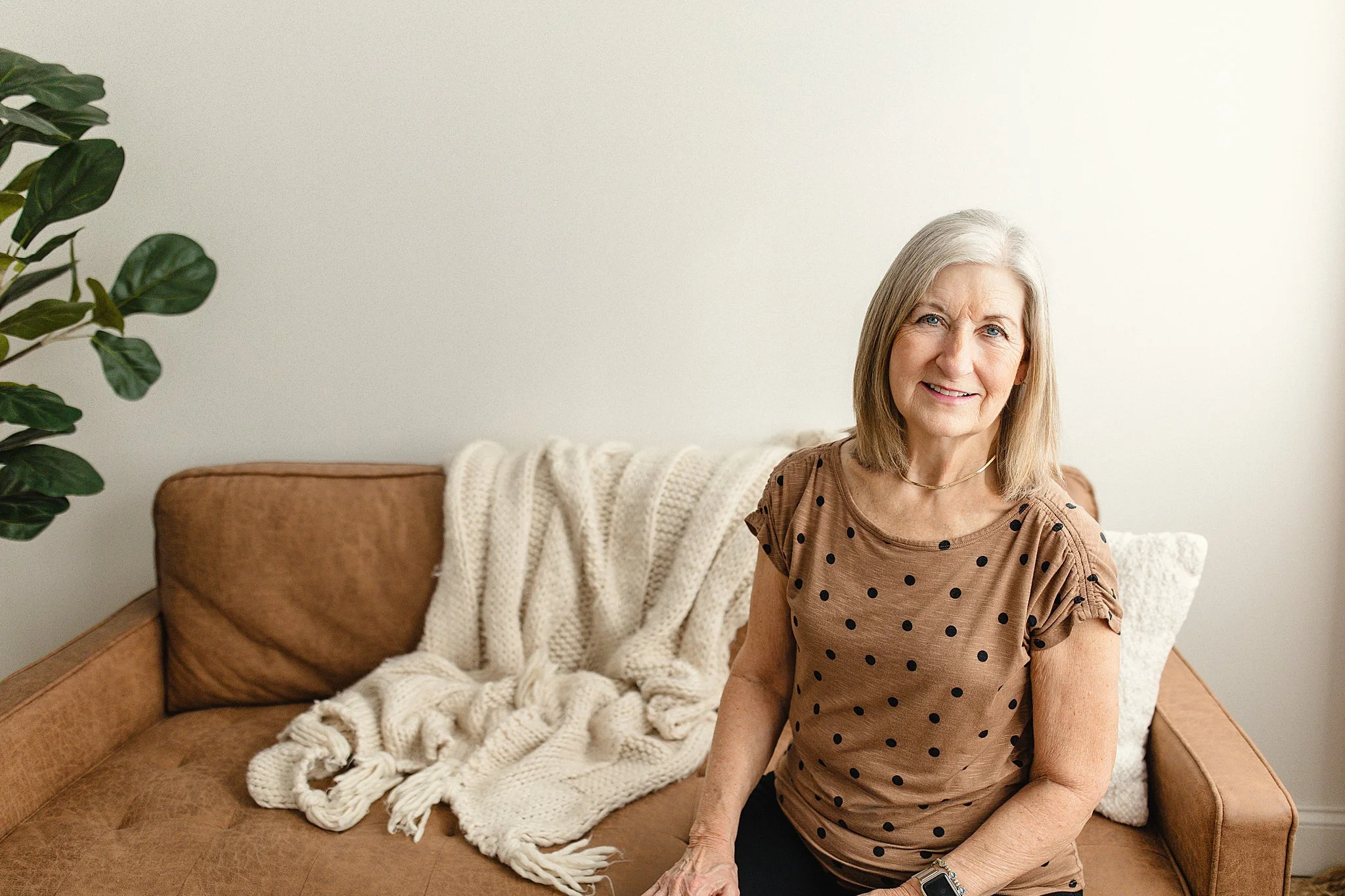An elderly woman with gray hair sitting on a tan couch, wearing a brown polka-dot shirt, smiling at the camera in a cozy living room for headshots in Naperville, IL.
