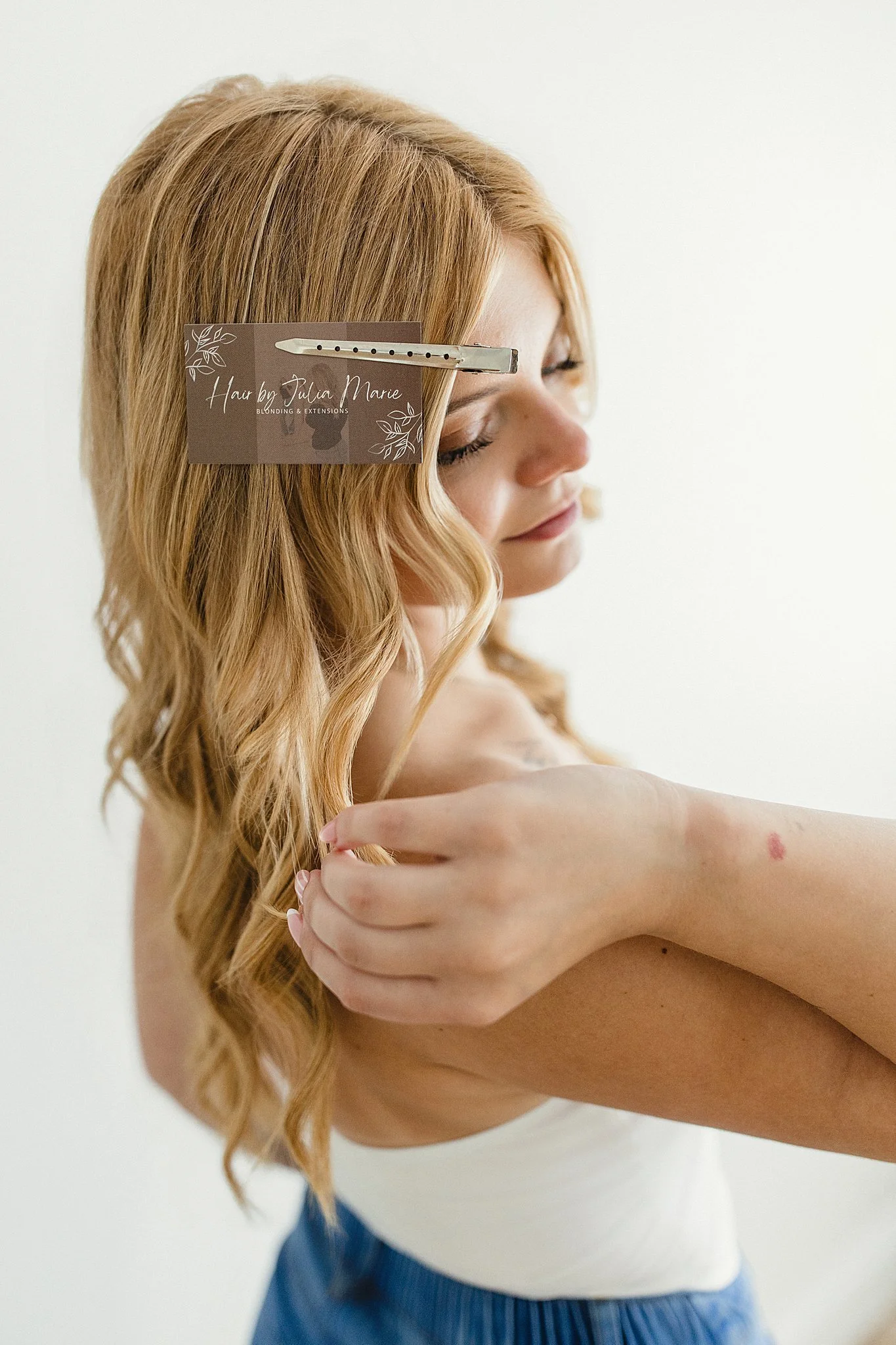 A woman with long, wavy, blonde hair is holding her arm across her chest, displaying a red mark or bruise on her forearm. She is wearing a white sleeveless top and blue pants, with her eyes closed for small business branding photography.