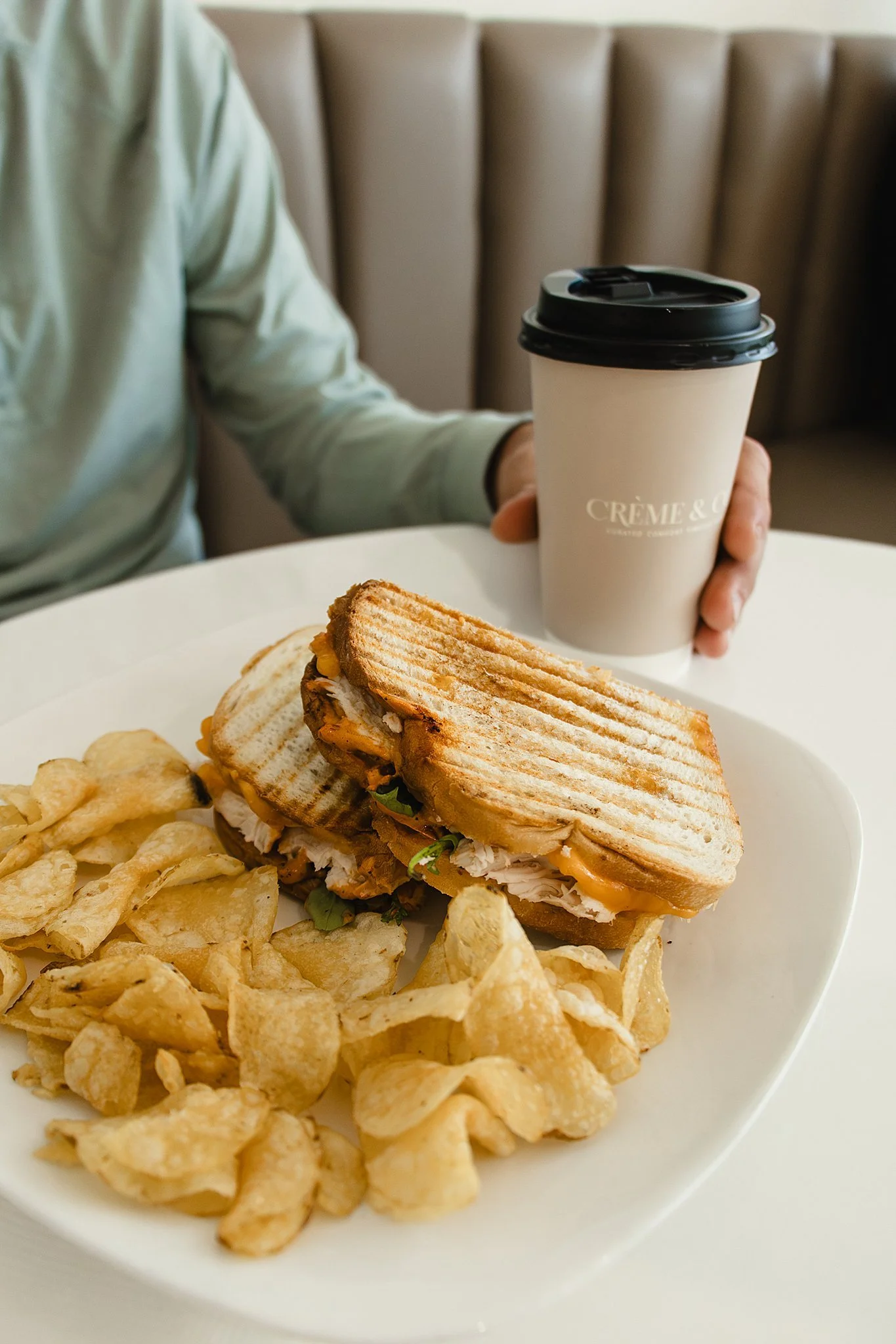 A plate of potato chips, a chicken sandwich with lettuce and cheese, and a partially visible pickle slices, with a person holding a coffee cup in the background at a dining table with brand photographers near me.