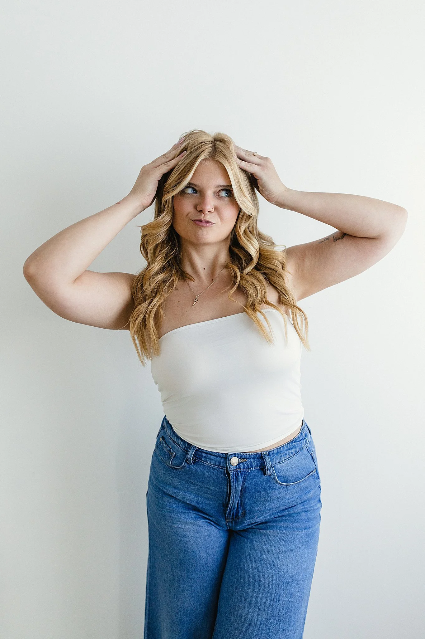 A young woman with long blonde hair and blue eyes, wearing a white strapless top and high-waisted blue jeans, standing against a plain white wall, with her hands on her head, looking slightly to her left for branding photos near me.