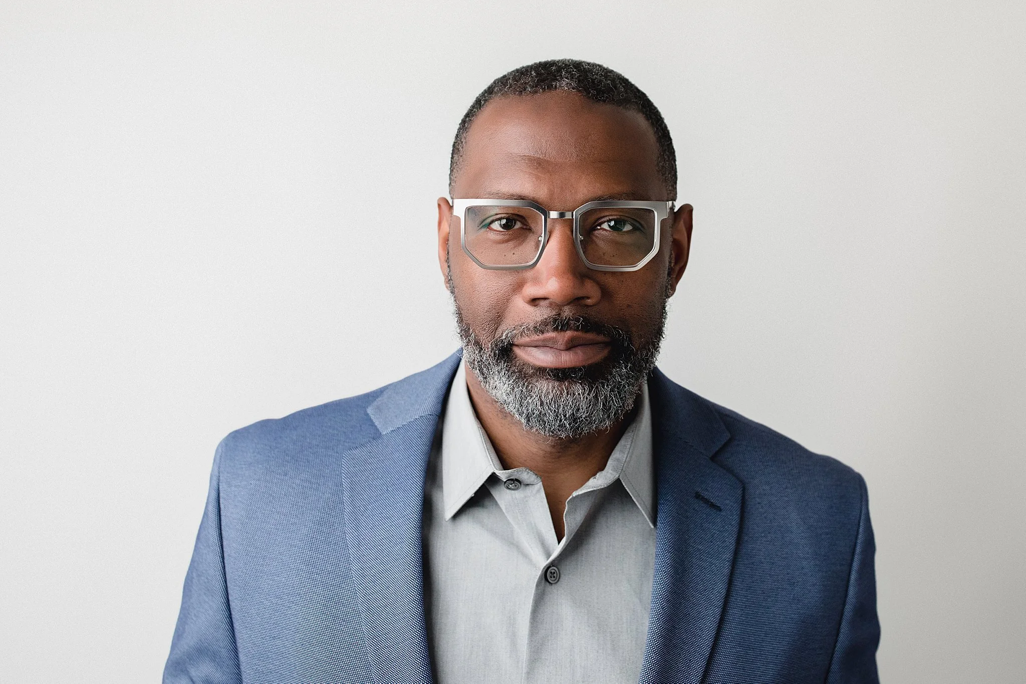 A confident middle-aged African American man in a light blue suit and light gray shirt, wearing glasses, standing against a plain white background with Naperville headshot photographer Marie Lopez of Ally and B Photography.